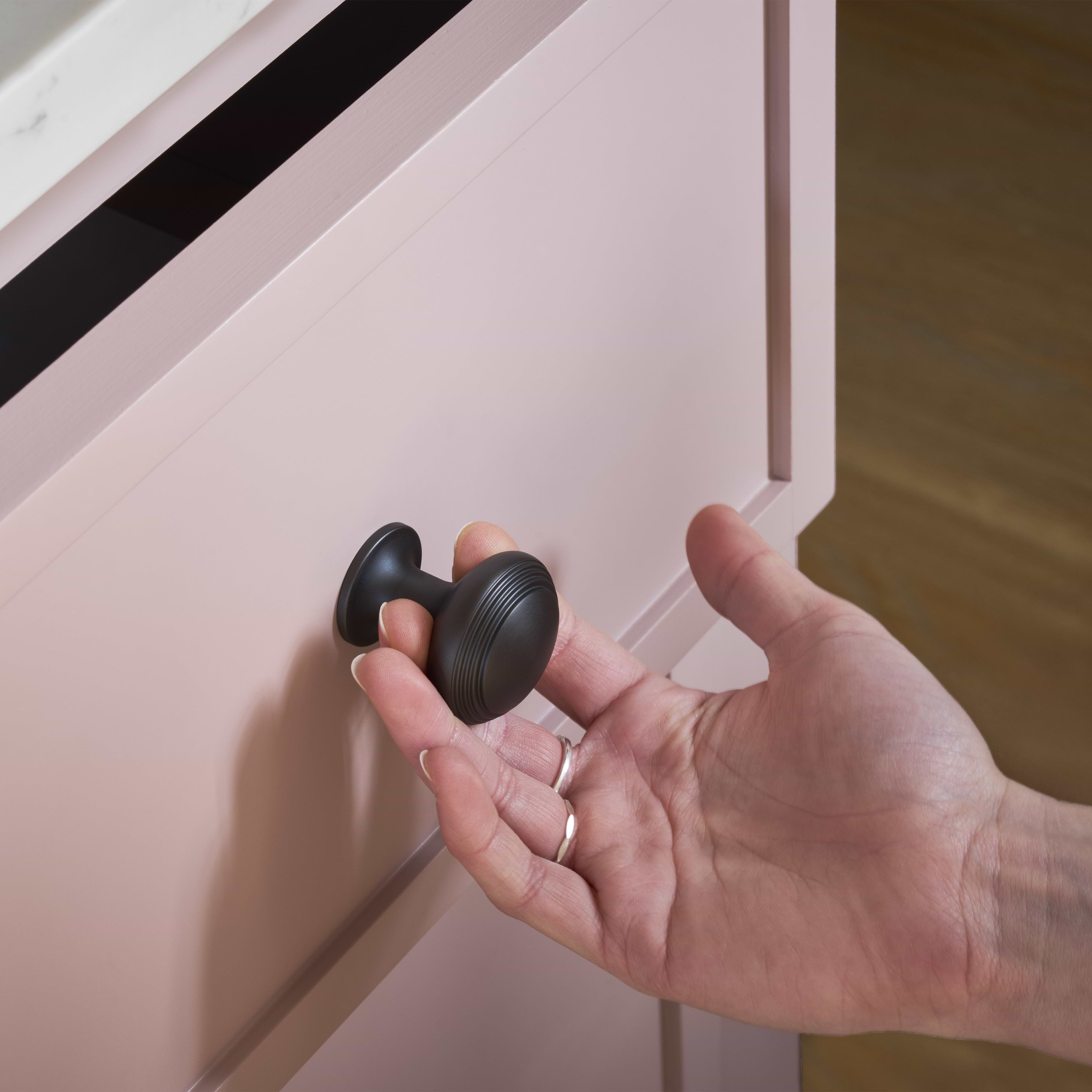 A hand grasps a bronze Barlow Furniture Knob Medium mounted on a light pink cabinet drawer.