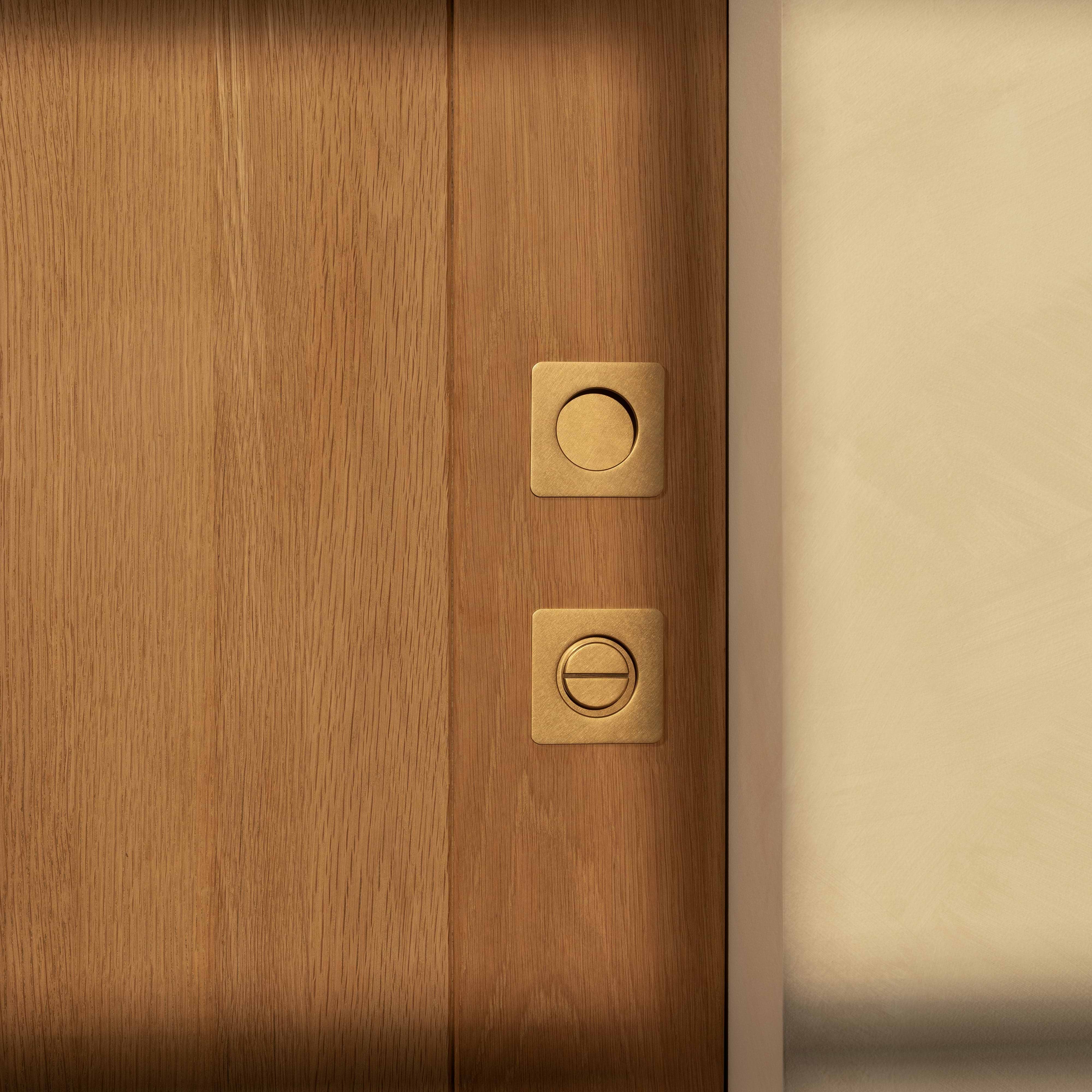 Close-up of a Regent Recessed Thumbturn in Antique Brass fitted on a wooden door, partially framed by a beige wall.