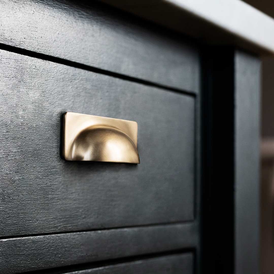 Close-up of an Elm Cup Handle Small in antique brass fitted to a dark green wooden drawer.