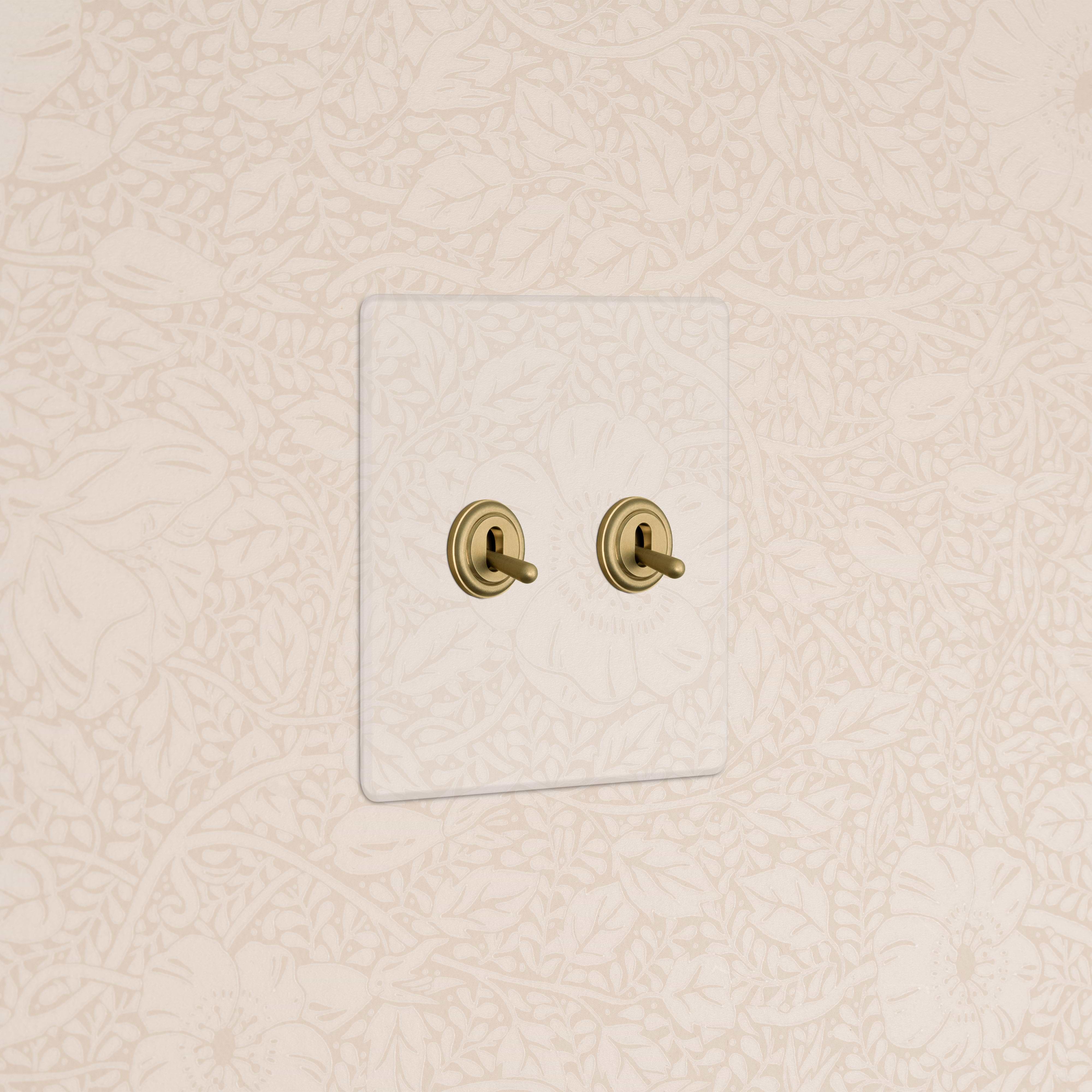 Kew 2G Toggle Switch in clear antique brass with two brass toggles, mounted on a floral-patterned wall.