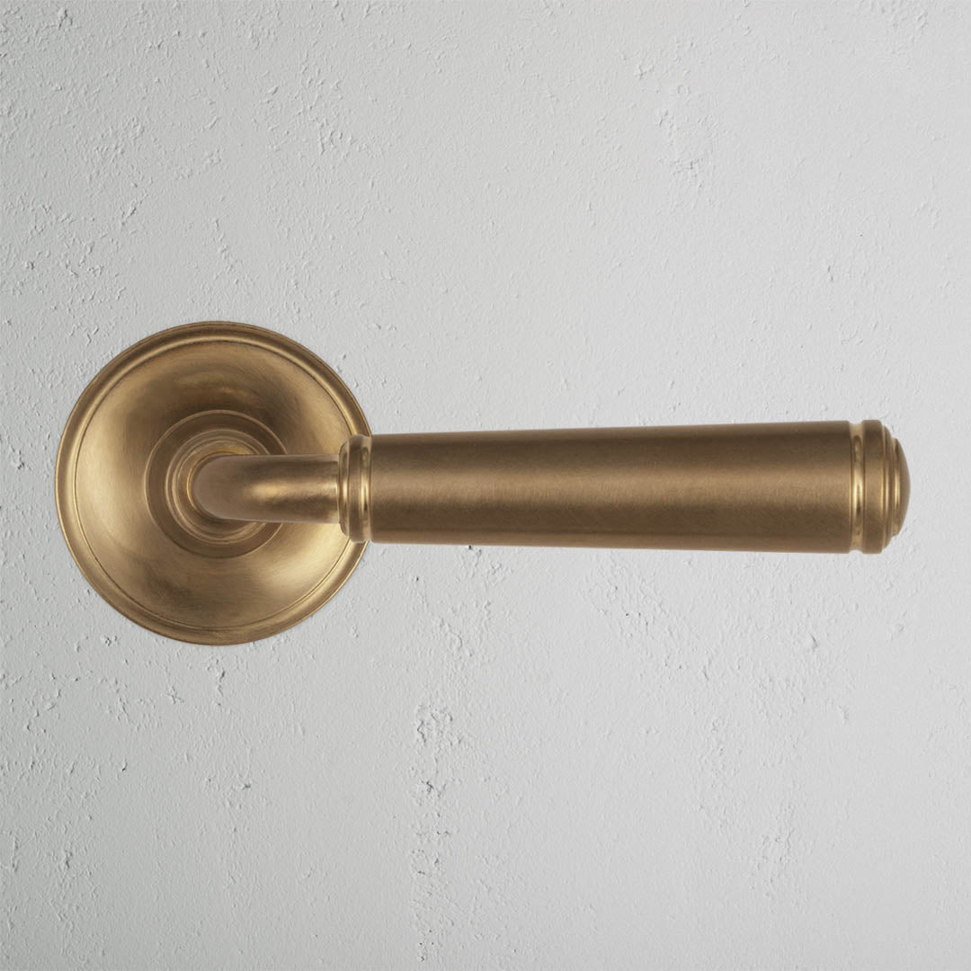 Digby Passage Door Handle Set in Antique Brass with a round base, shown installed on a white textured wall.