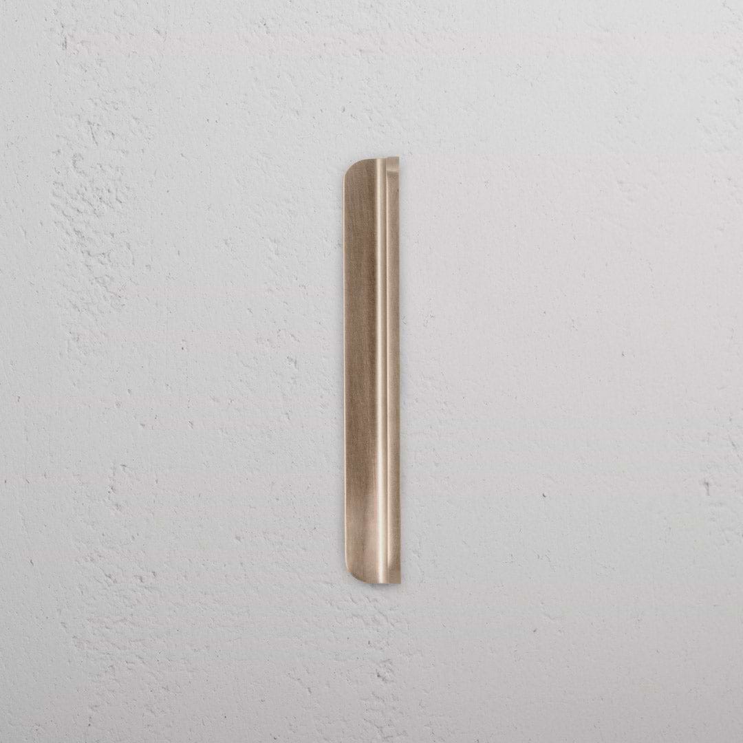 Oxford Furniture Handle Small in Antique Brass, slender and vertical, mounted on a textured white wall.