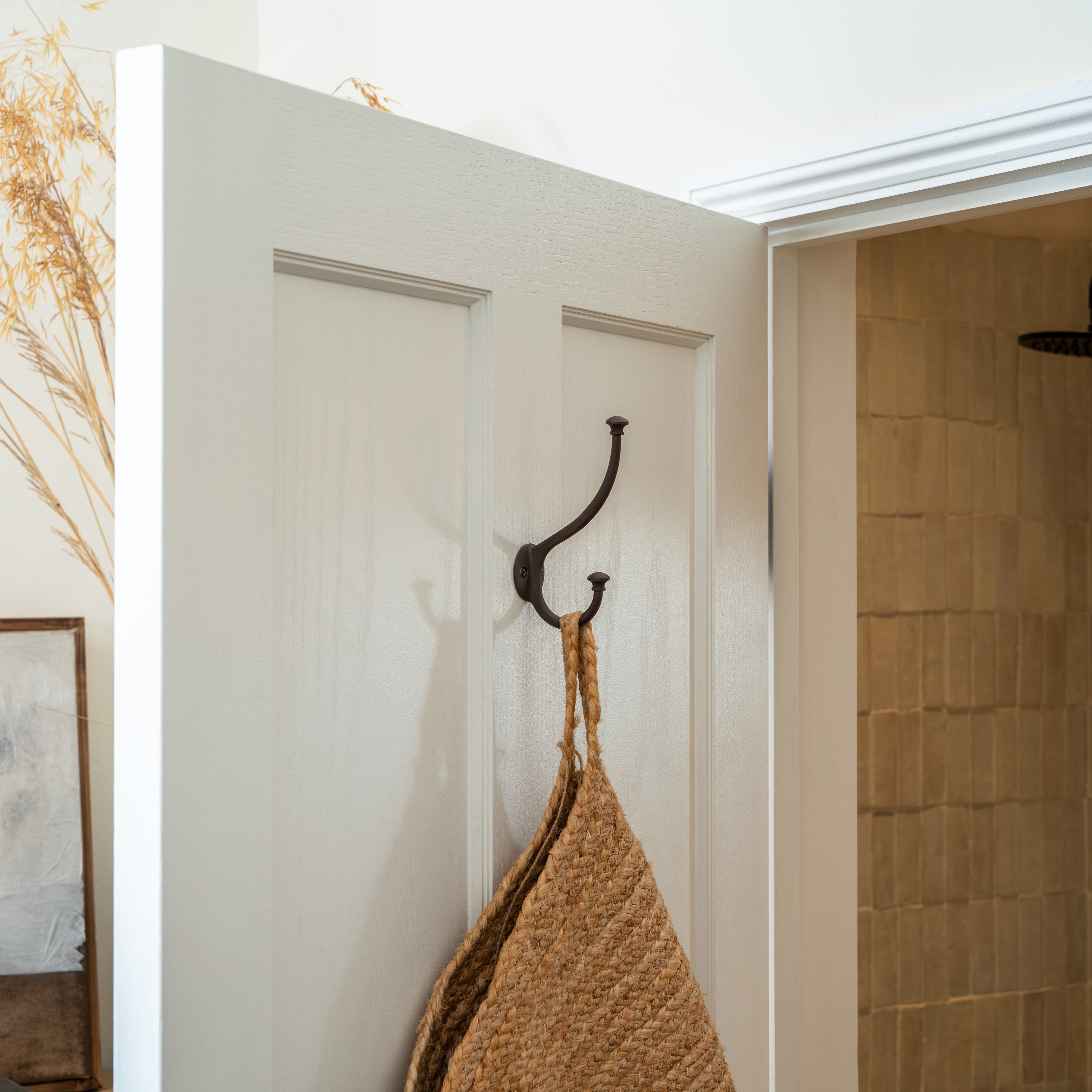 Bronze Mews Hook mounted on a white door, holding a tan woven bag in a softly lit bathroom setting.