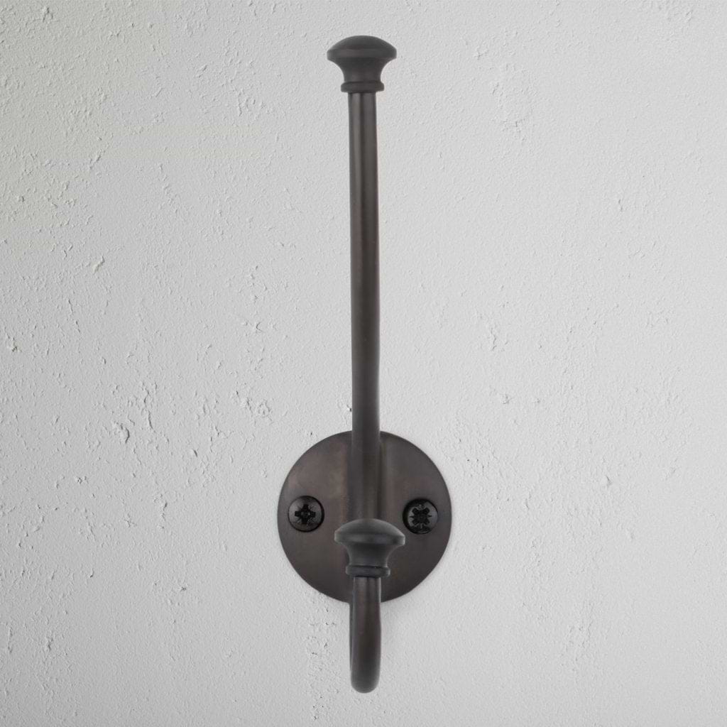 Bronze Mews Hook with round base, two screws, and dual arms mounted on a white textured wall.