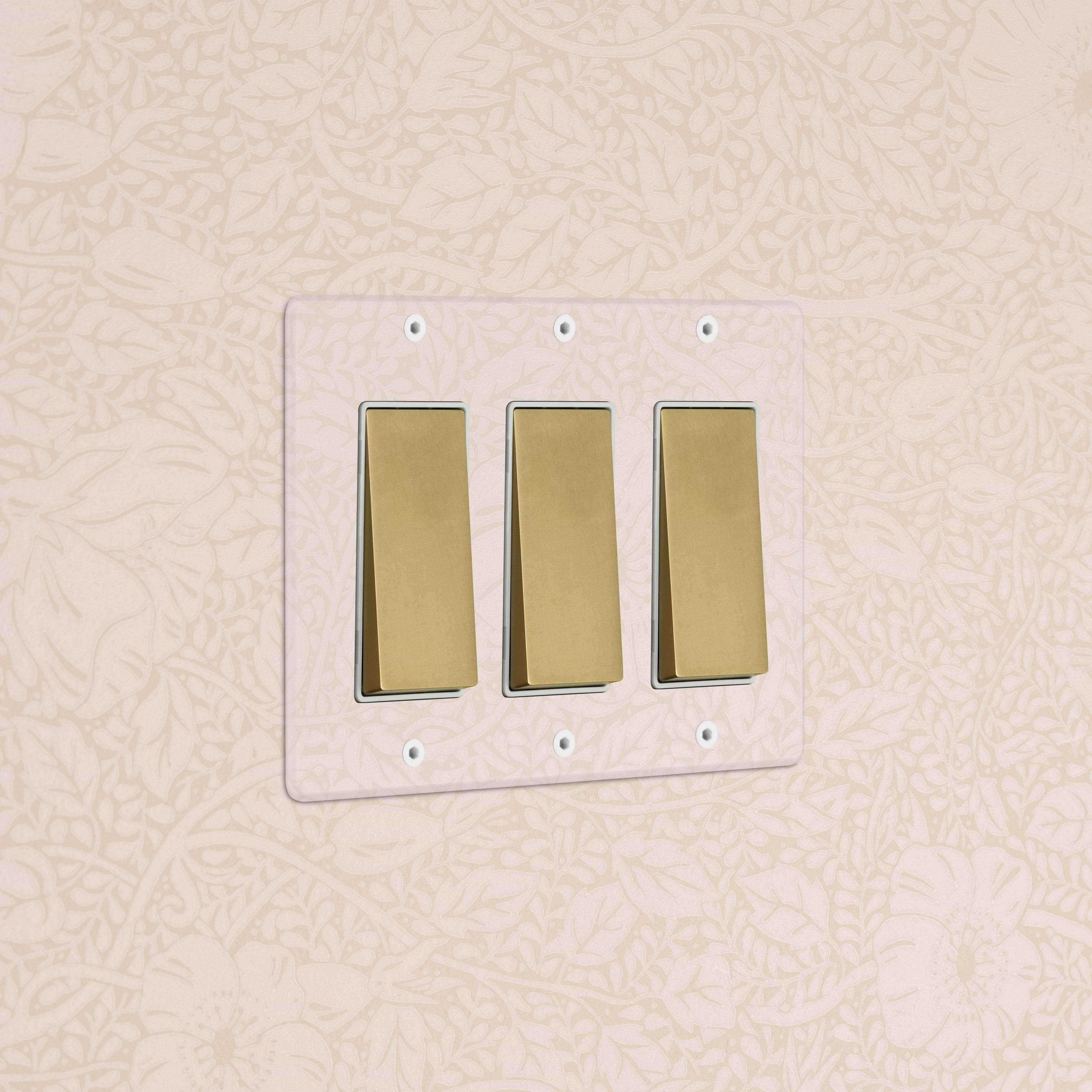Kew 3G Rocker Switch in clear antique brass white, wall-mounted against beige floral-patterned wallpaper.