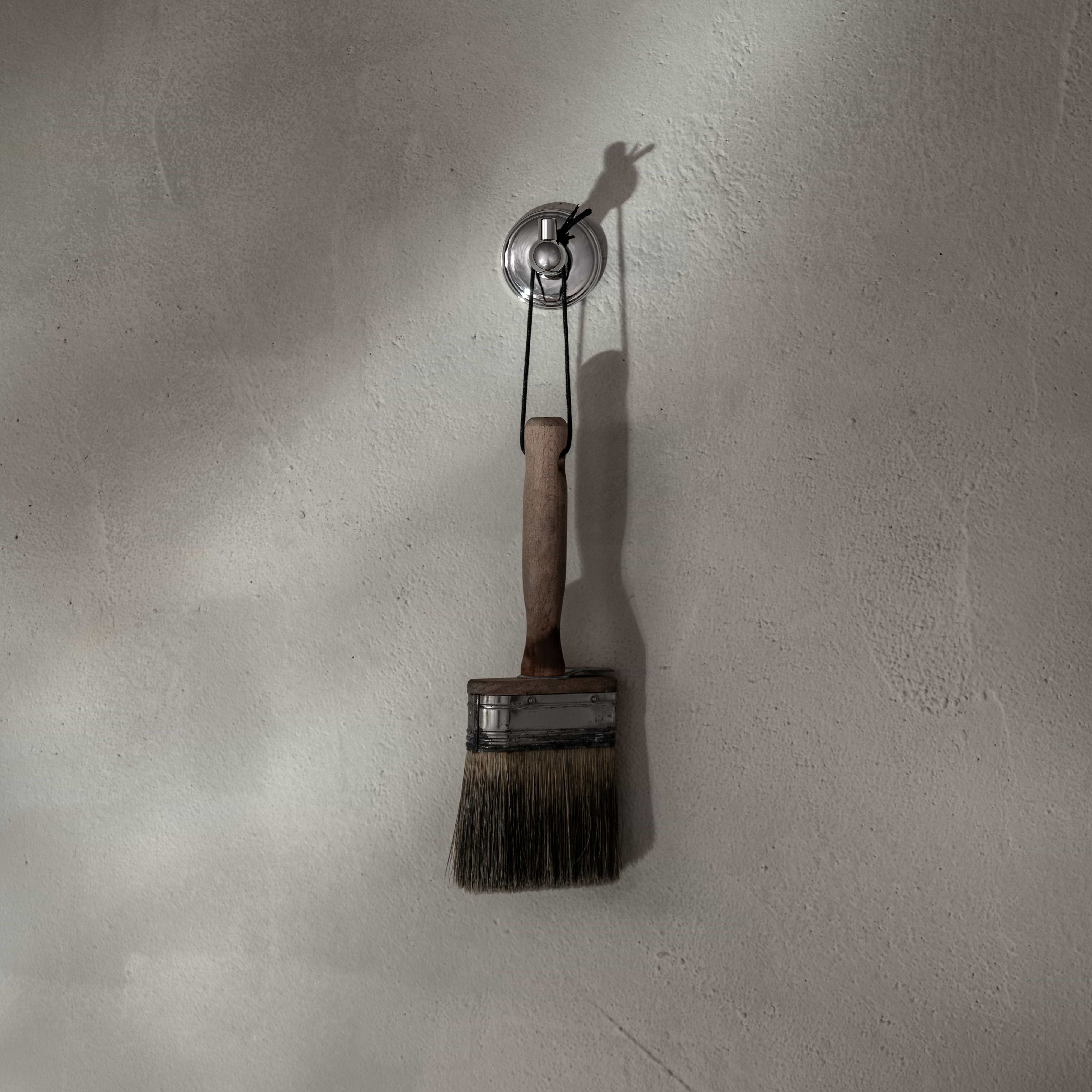 Polished nickel Hyde Hook mounted on a light-colored wall, holding a large paintbrush by its handle.