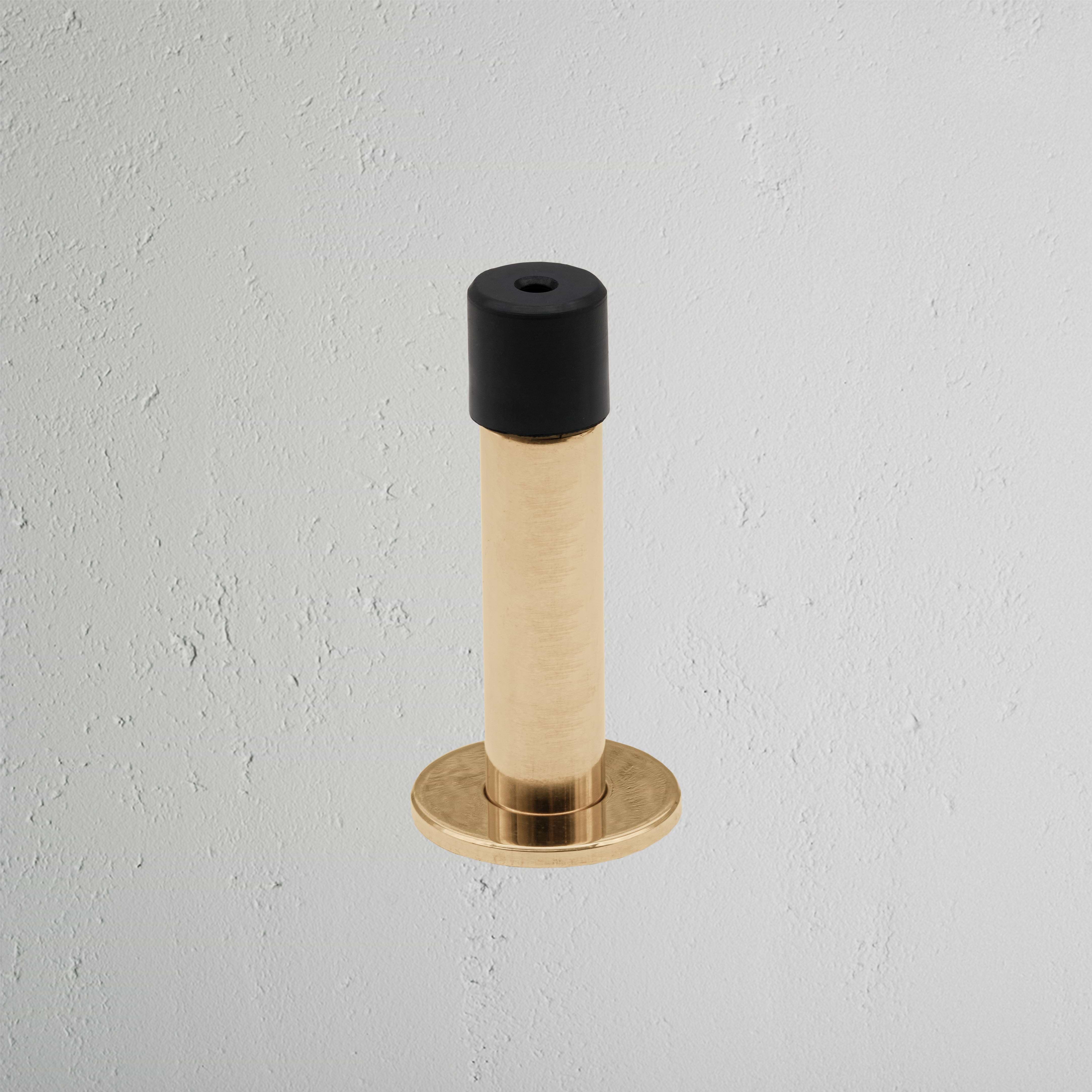 Montpelier Door Stop in antique brass finish with black rubber tip, mounted on a light gray wall.