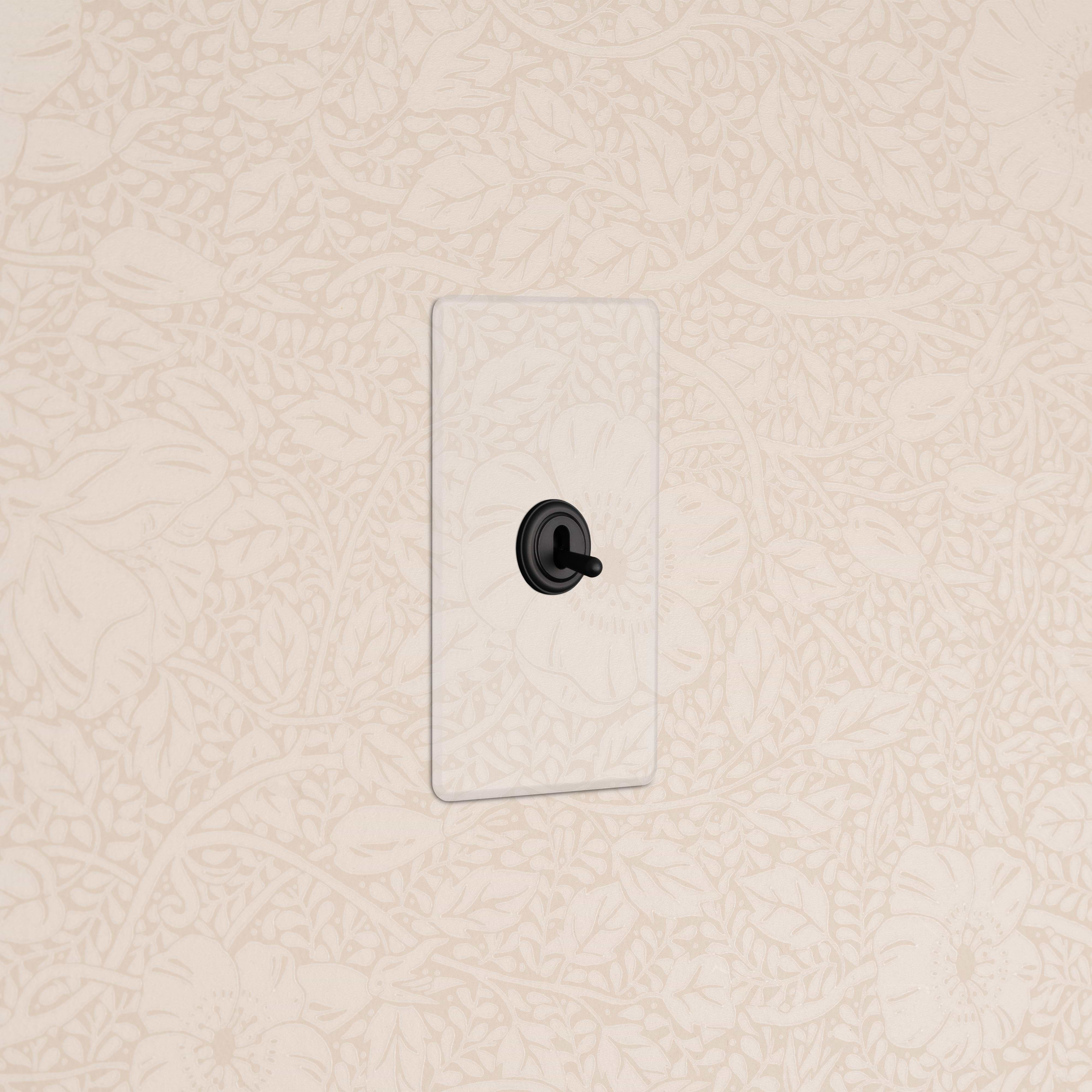 Kew 1G Four Way Toggle Switch in clear bronze finish, shown installed on a patterned beige wall.
