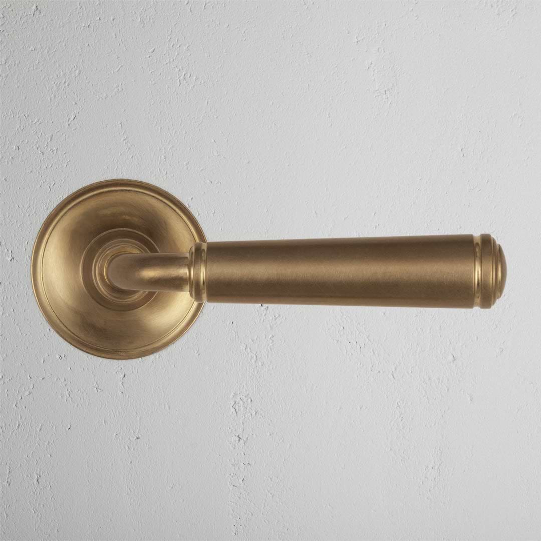 Digby Passage Door Handle Set in Antique Brass with a round base, shown installed on a white textured wall.
