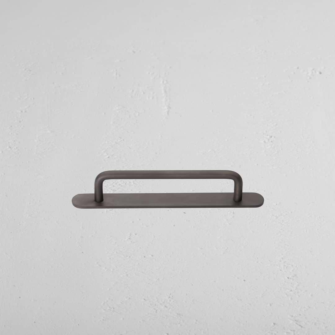 Small bronze Kilburn furniture handle with rounded ends, mounted horizontally on a textured white wall.