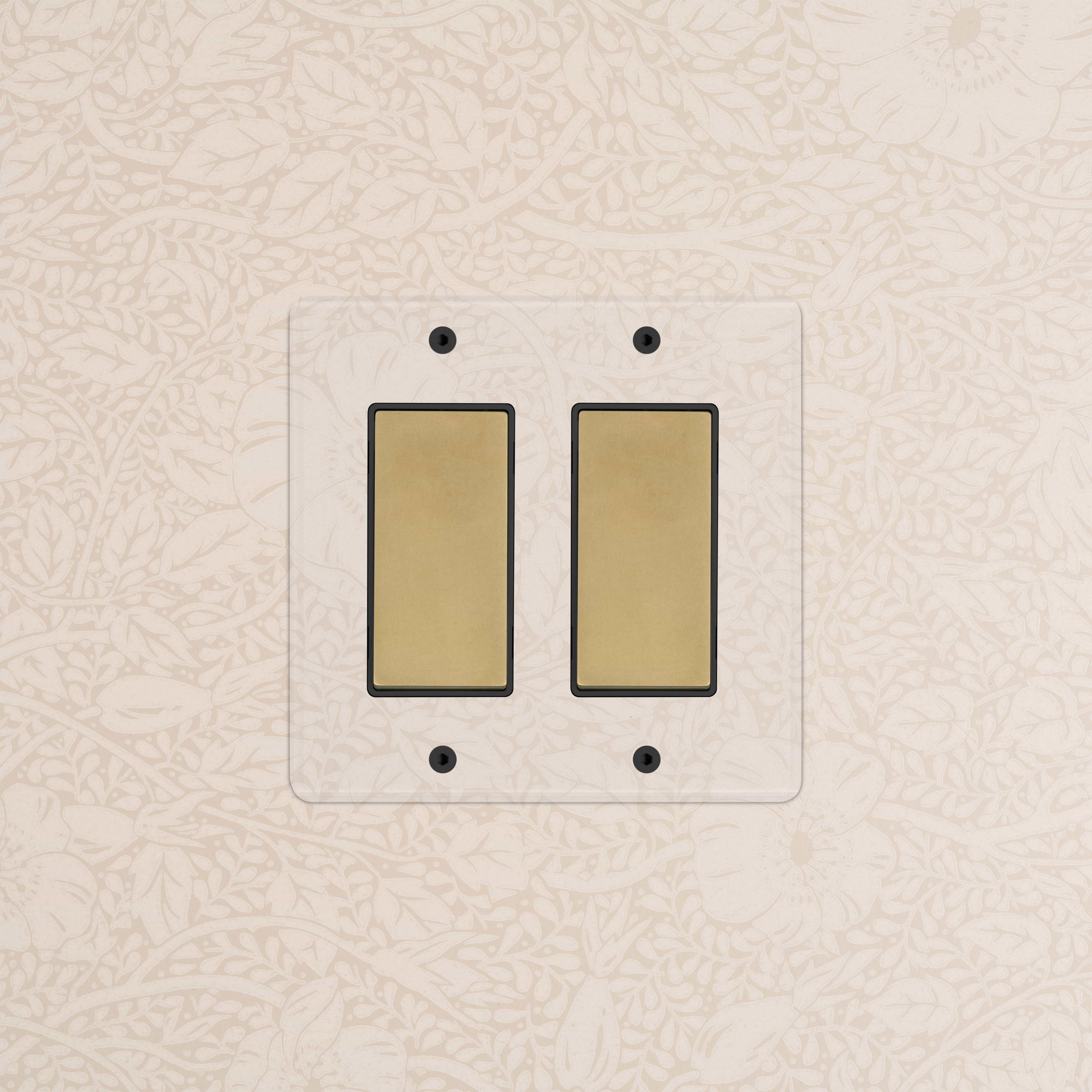 Kew 2G Rocker Switch in clear antique brass black, shown mounted on floral-patterned wallpaper.