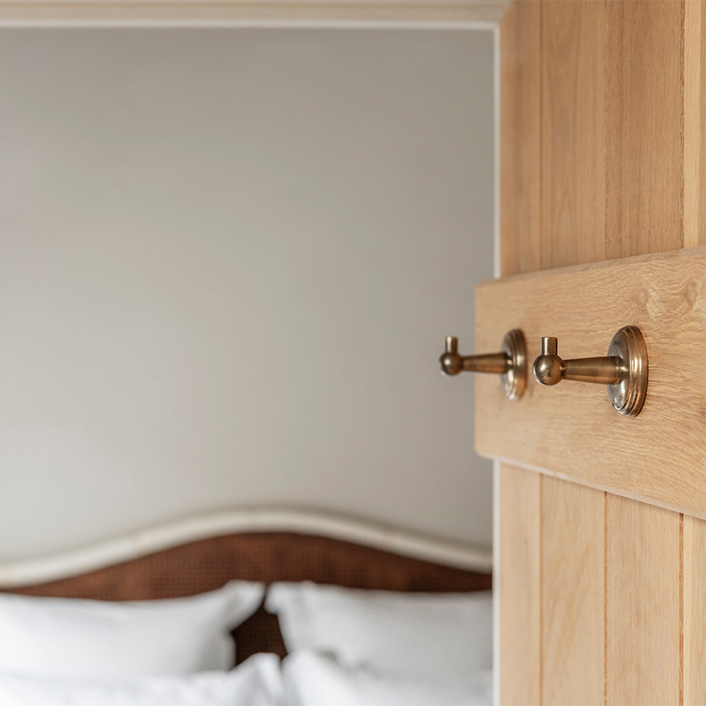 Two Hyde Hooks in antique brass mounted on a wooden door, bedroom with bed in background.