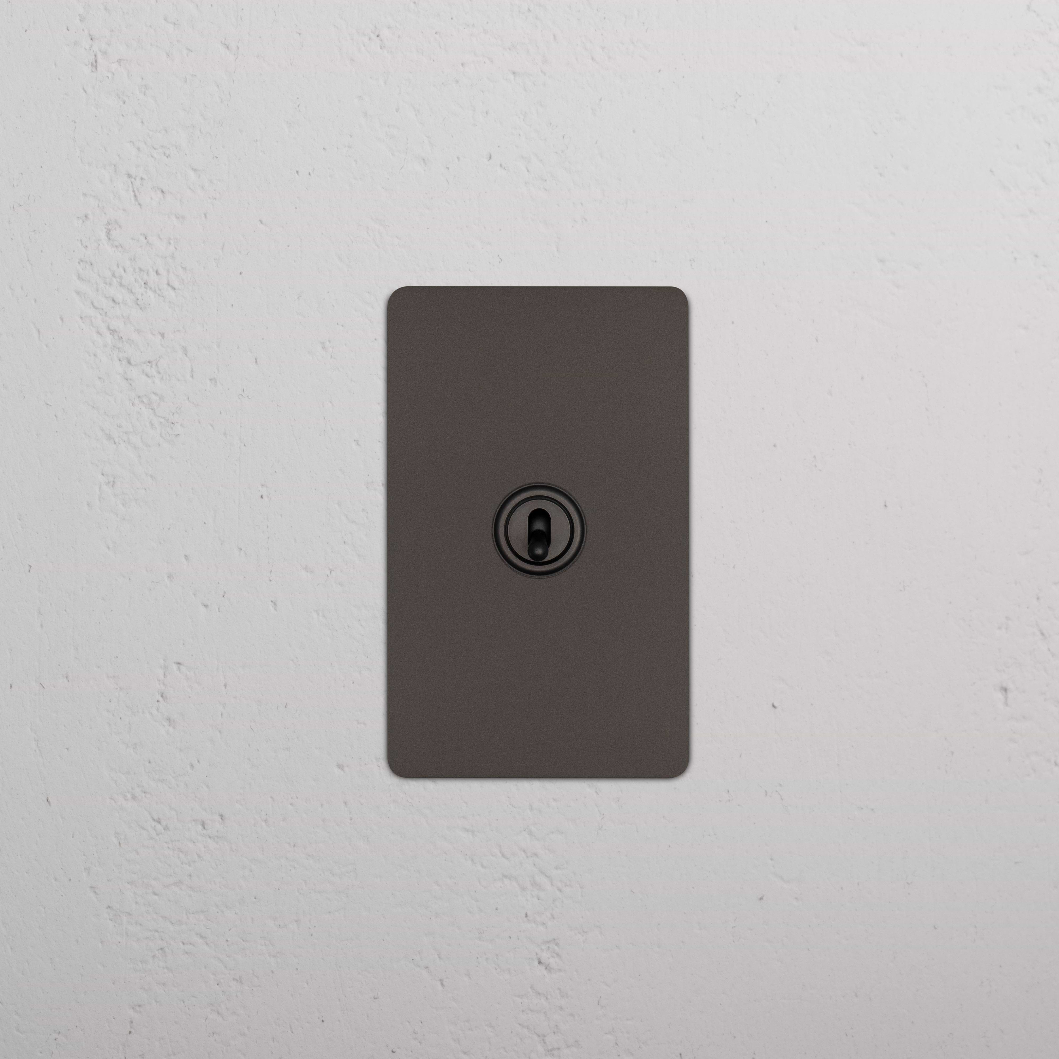 Bronze Kew 1G Toggle Switch with a central toggle on a rectangular plate, mounted on a white wall.