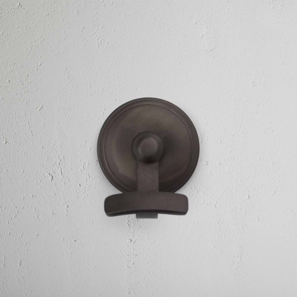 Portland Hook in dark bronze with a round base, featuring a modern design, mounted on a white wall.