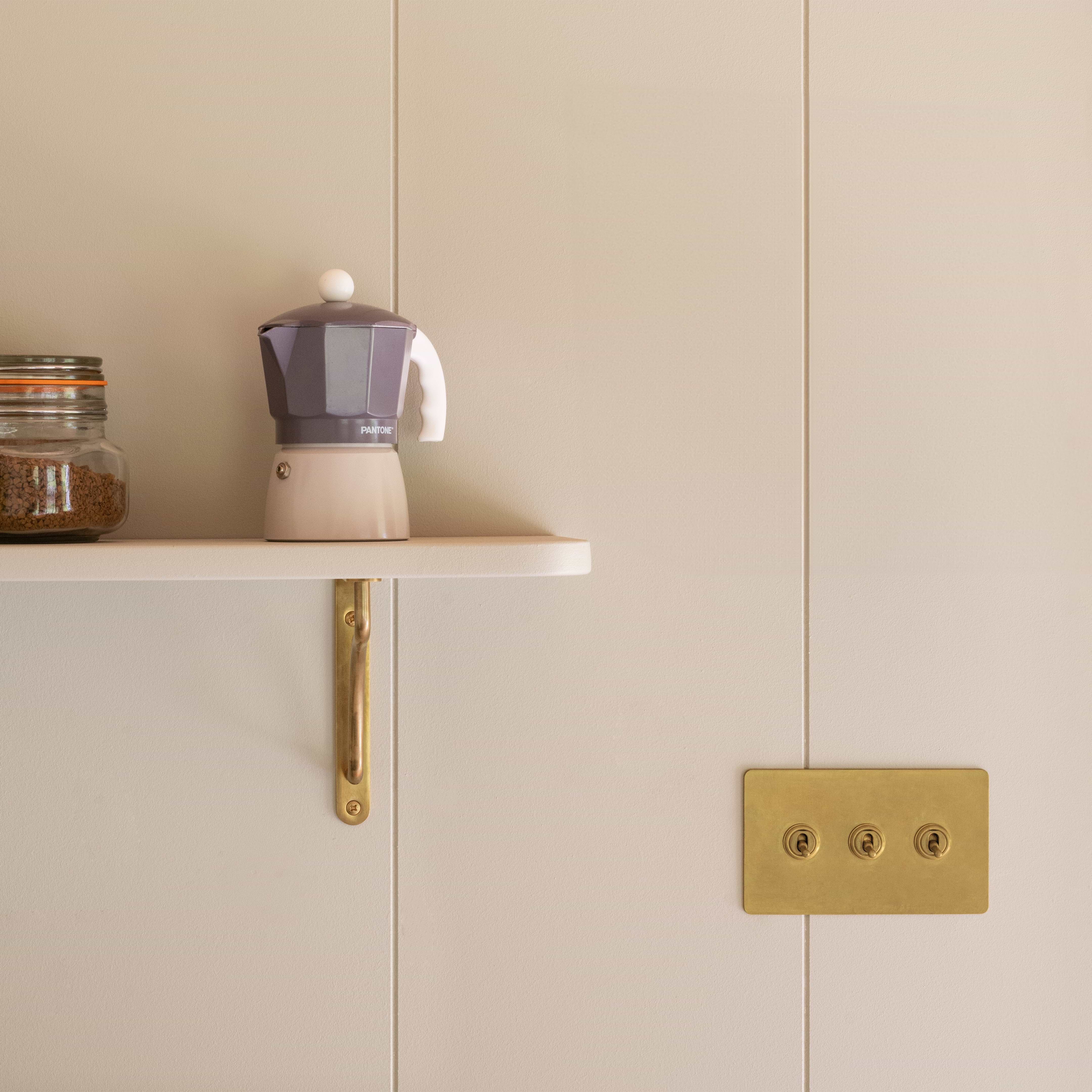A small coffee pot and jar on a shelf with Corston's Kilburn 200mm Antique Brass Bracket, beside a brass light switch on beige wall.