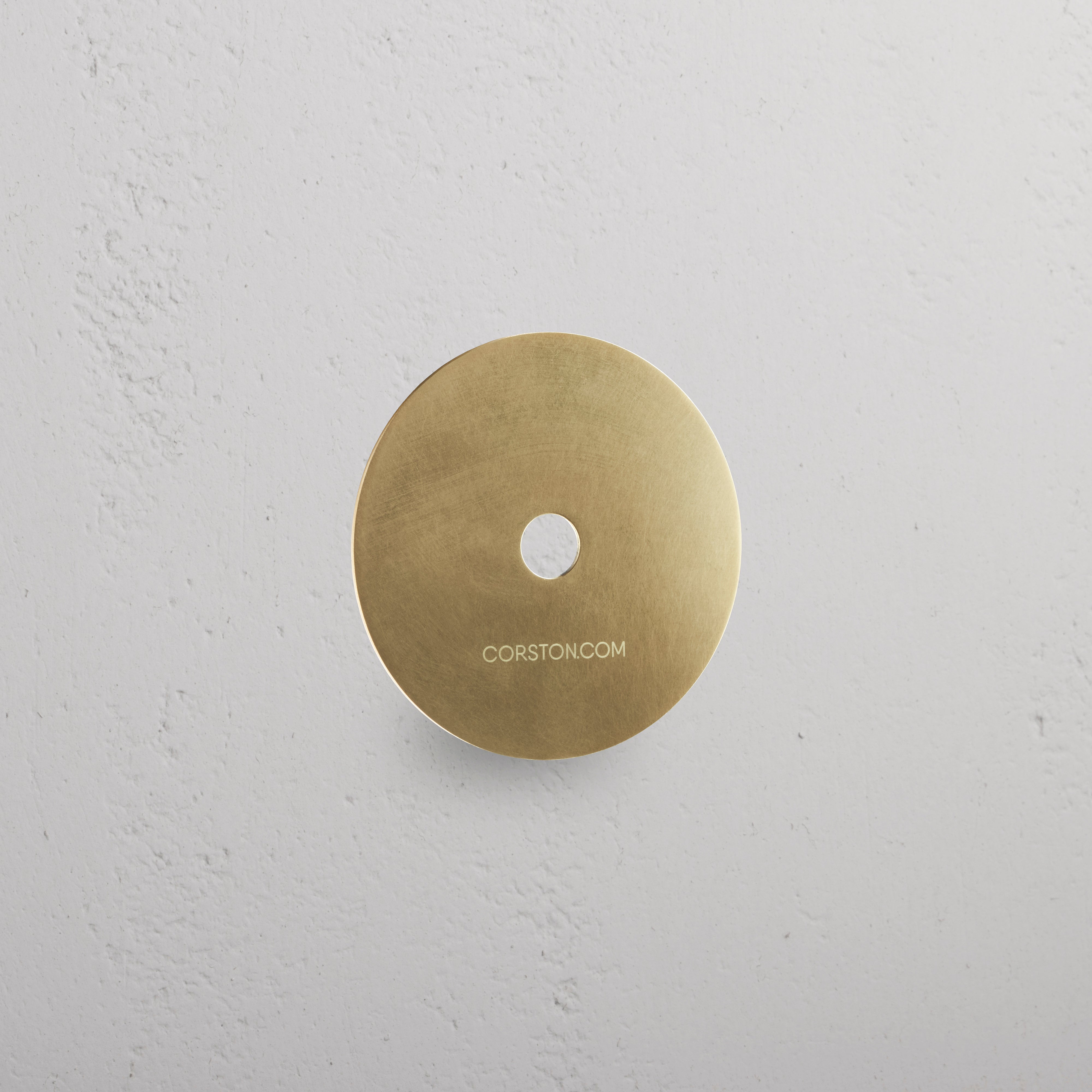 Circular Sample Finish in Antique Brass with central hole, mounted on white textured wall, labeled 'CORSTON.COM'.
