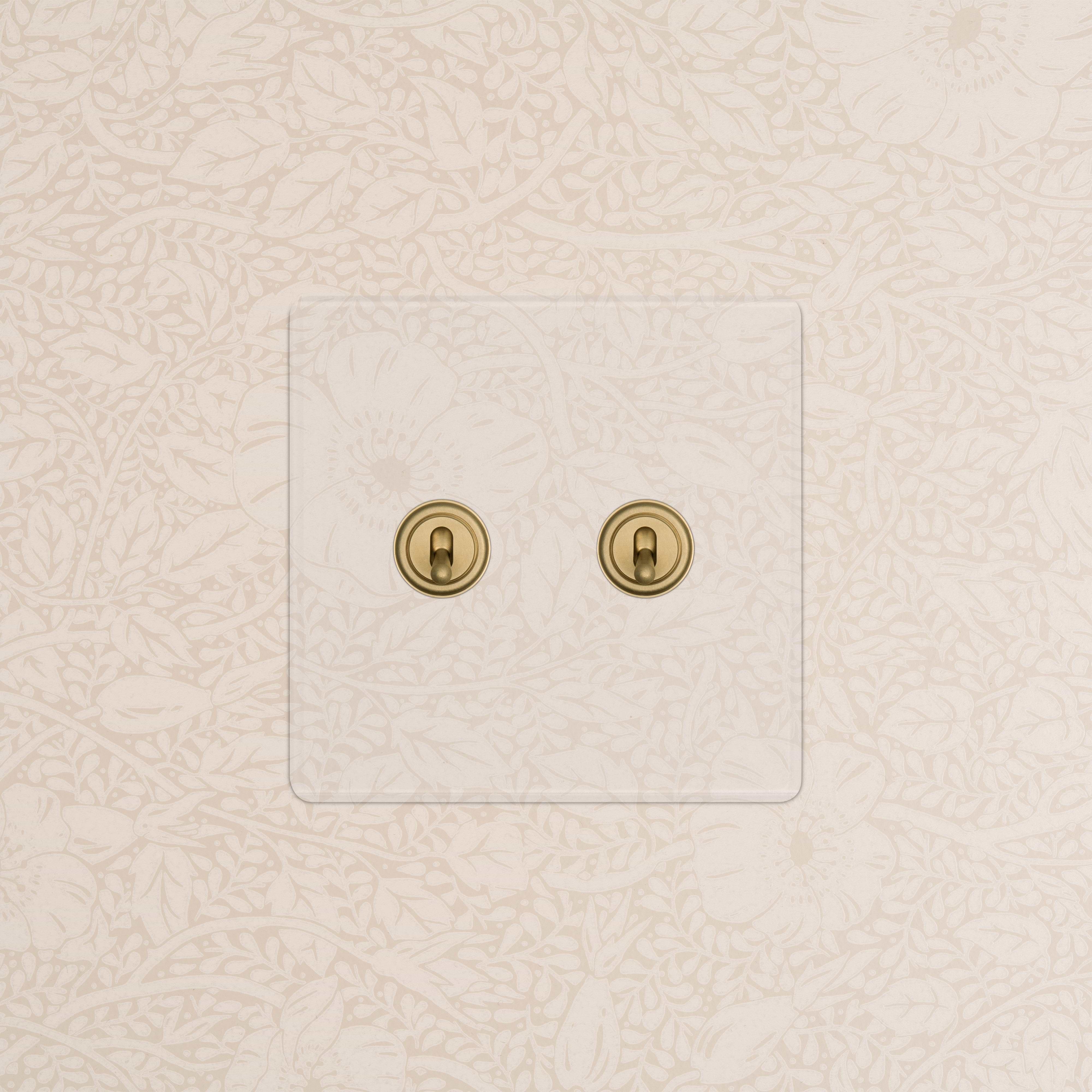 Kew 2G Toggle Switch in clear antique brass with elegant brass toggles on floral wallpaper background.