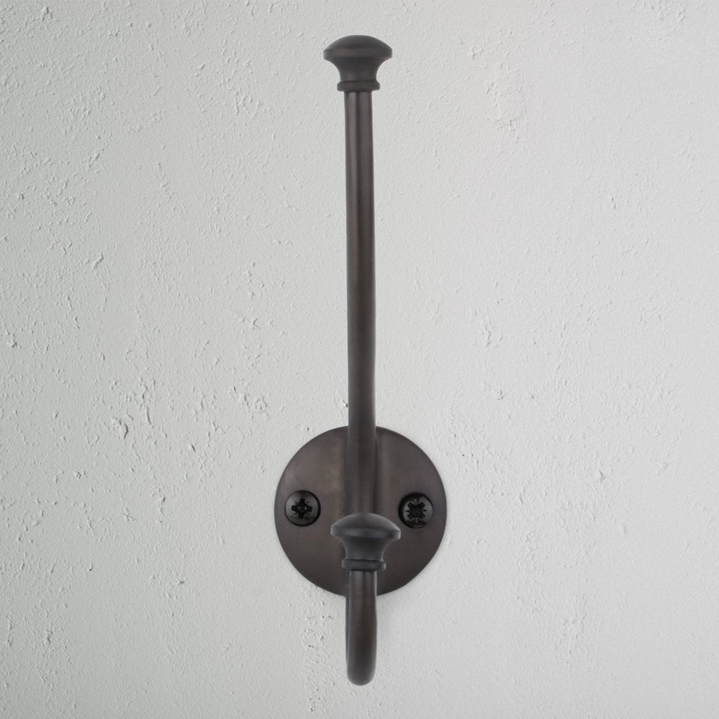 Bronze Mews Hook with round base, two screws, and dual arms mounted on a white textured wall.