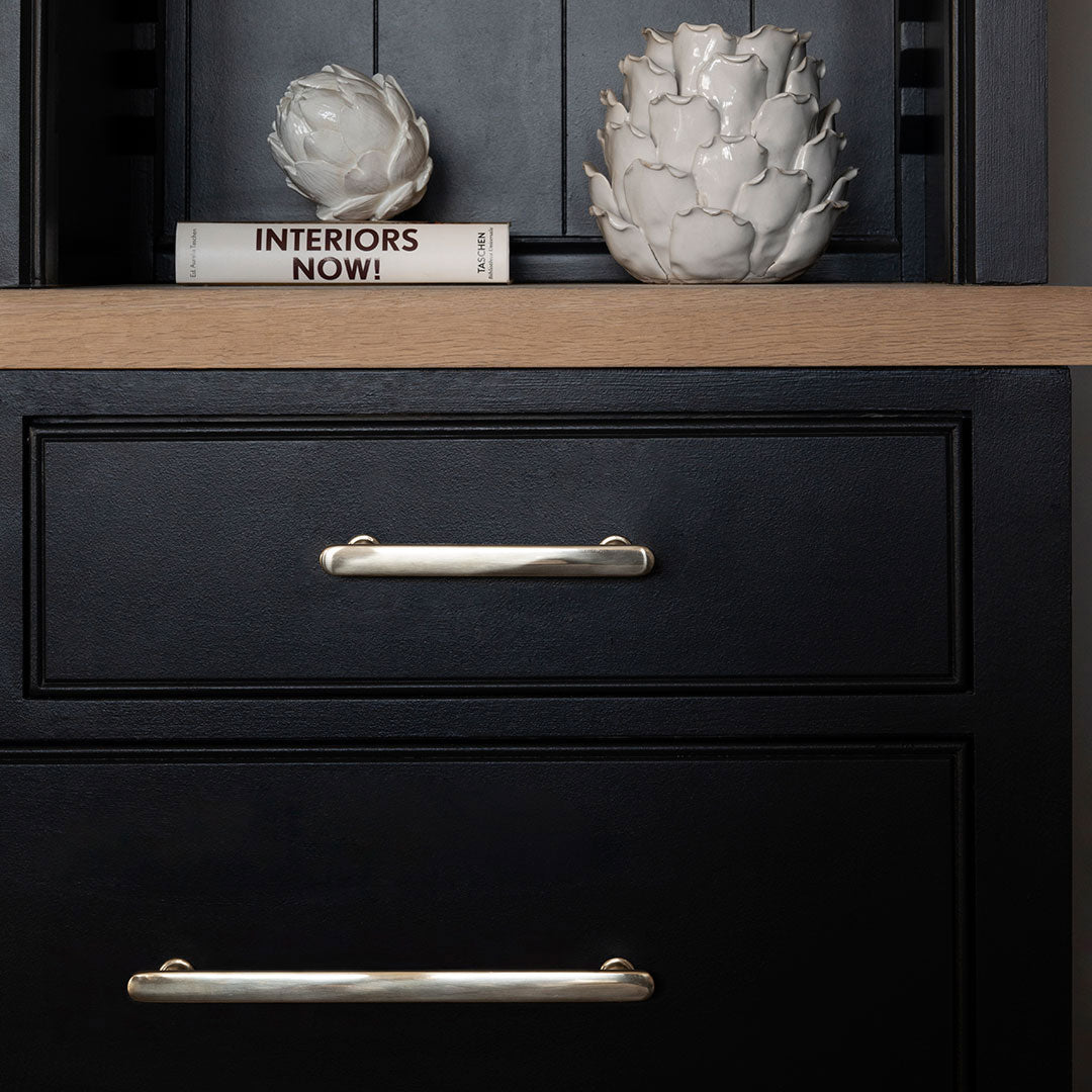 Medium polished nickel Milliner Furniture Handle mounted on a black cabinet with light wood surface and decor.