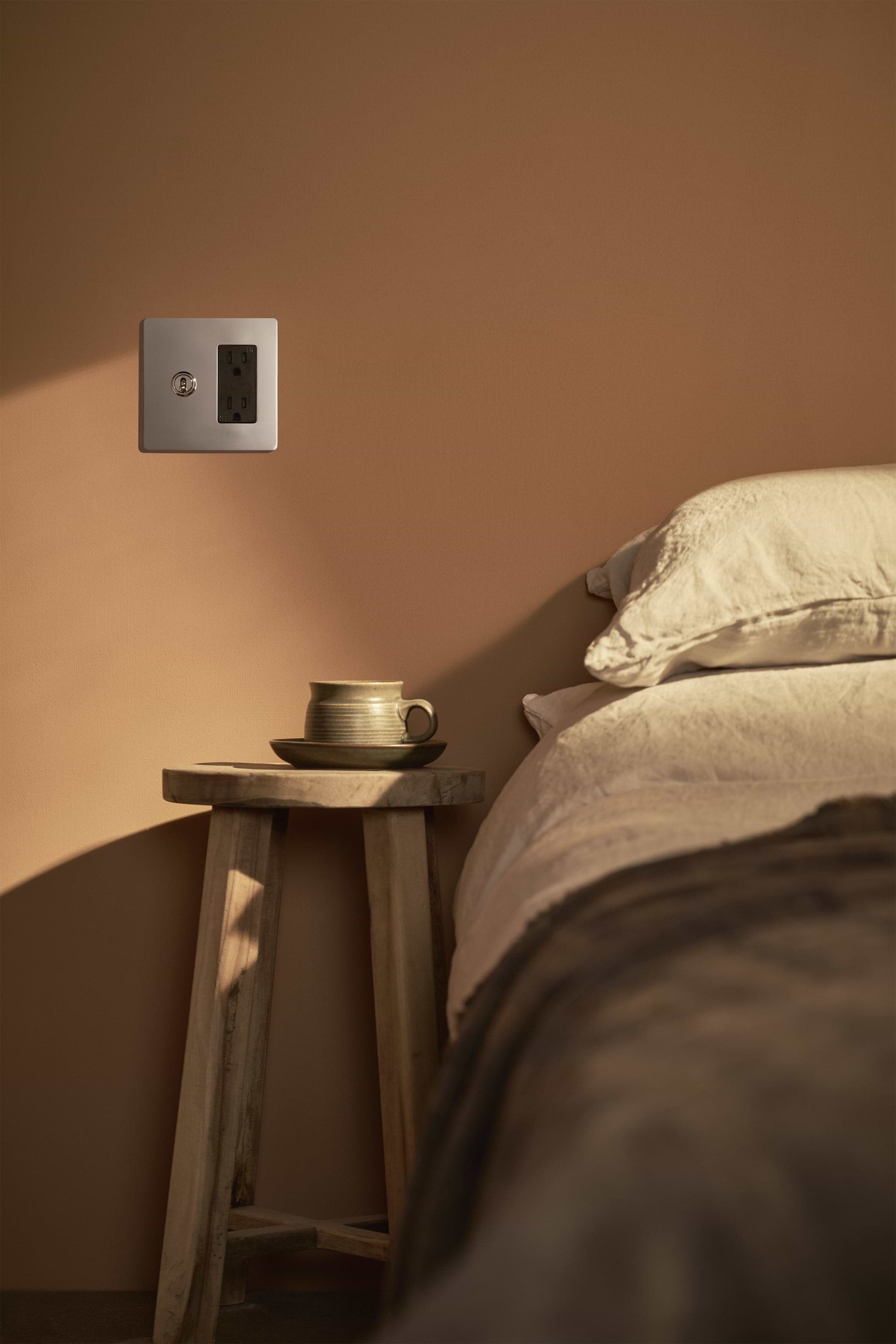 Kew 2G Toggle Switch & Outlet in Polished Nickel Black fitted on wall next to bed and bedside table.