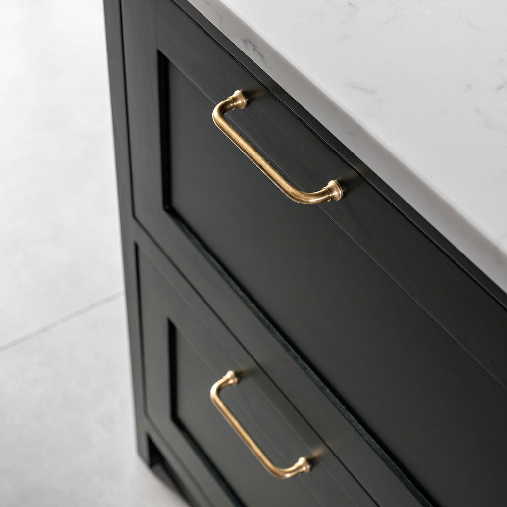 Small Antique Brass Sycamore Furniture Handle fitted on dark cabinet drawers beneath a white marble countertop.