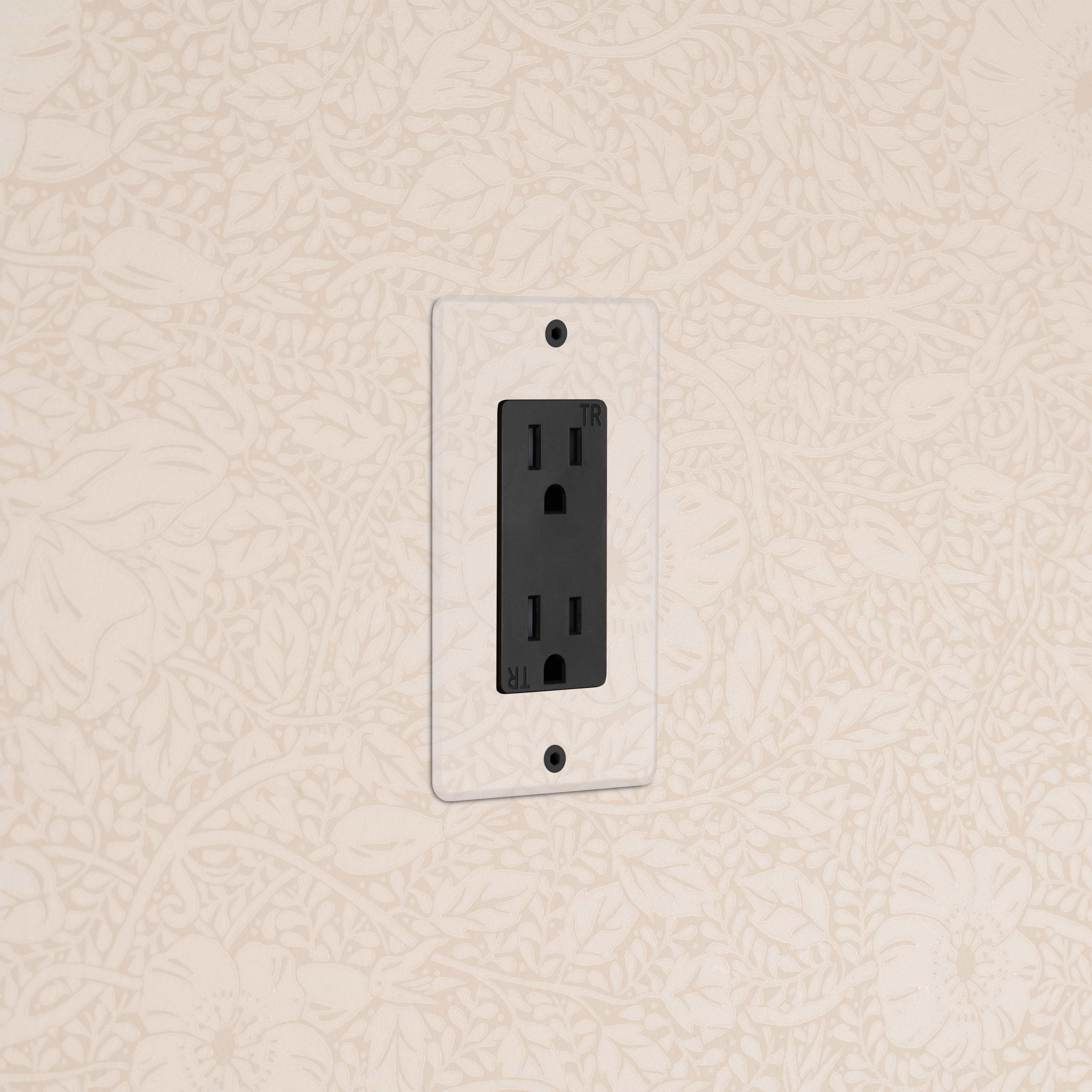 Kew 1G 15A electrical outlet in clear black finish, installed on a beige floral wallpapered wall.