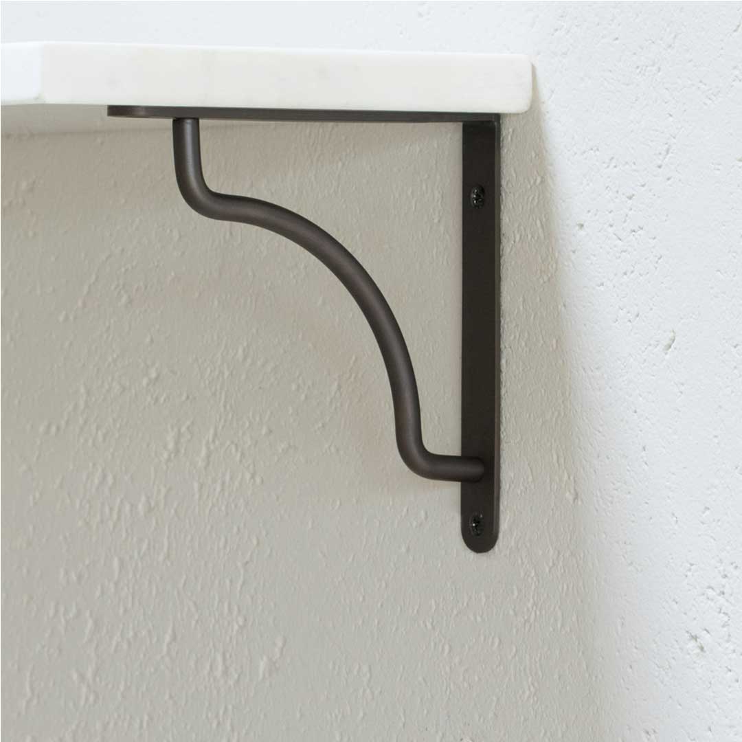 Kilburn Shelf Bracket Small in bronze, mounted on a white wall, supporting a white shelf with a curved support bar.