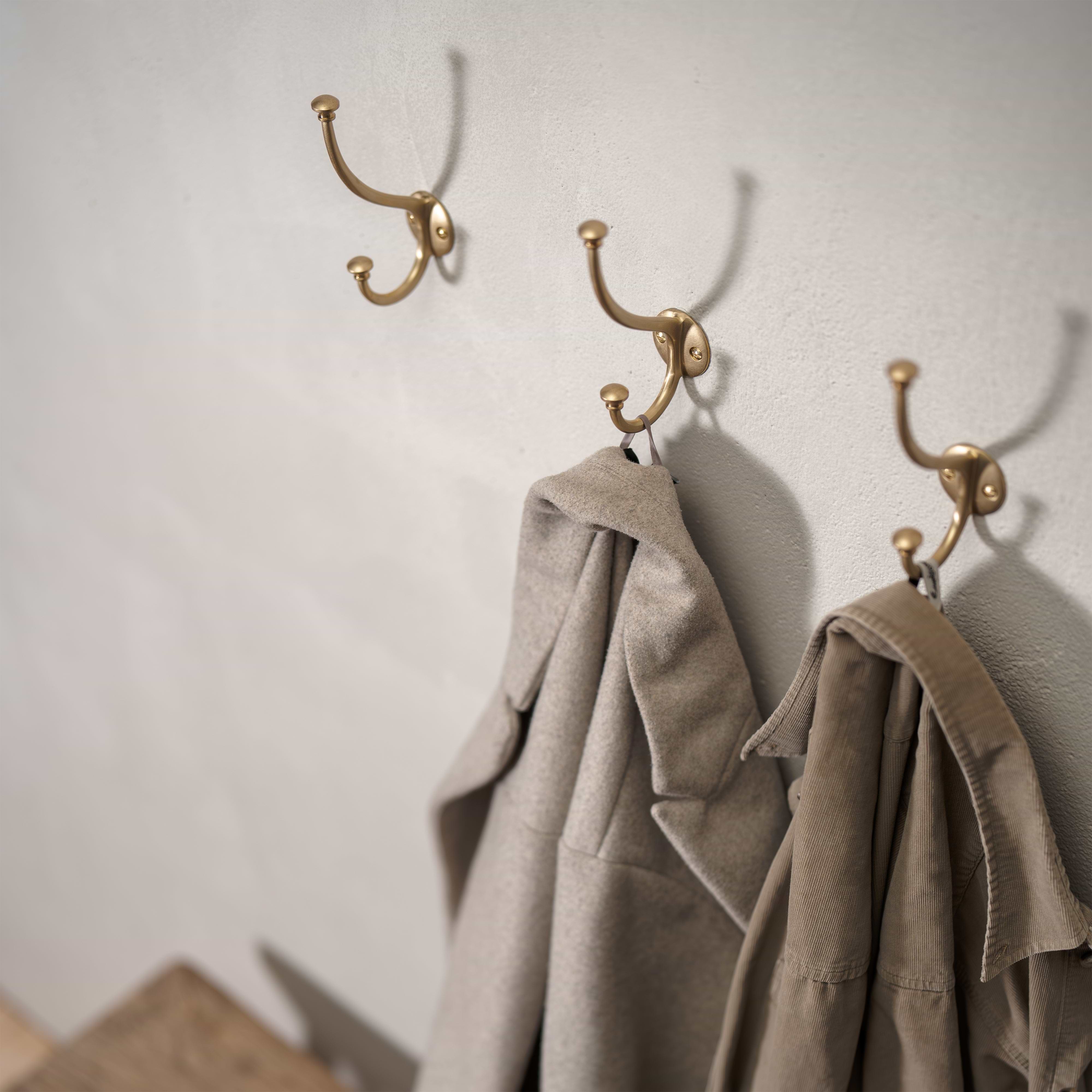 Three Mews Hook - Antique Brass wall hooks, two holding beige coats, mounted on a light-colored wall.