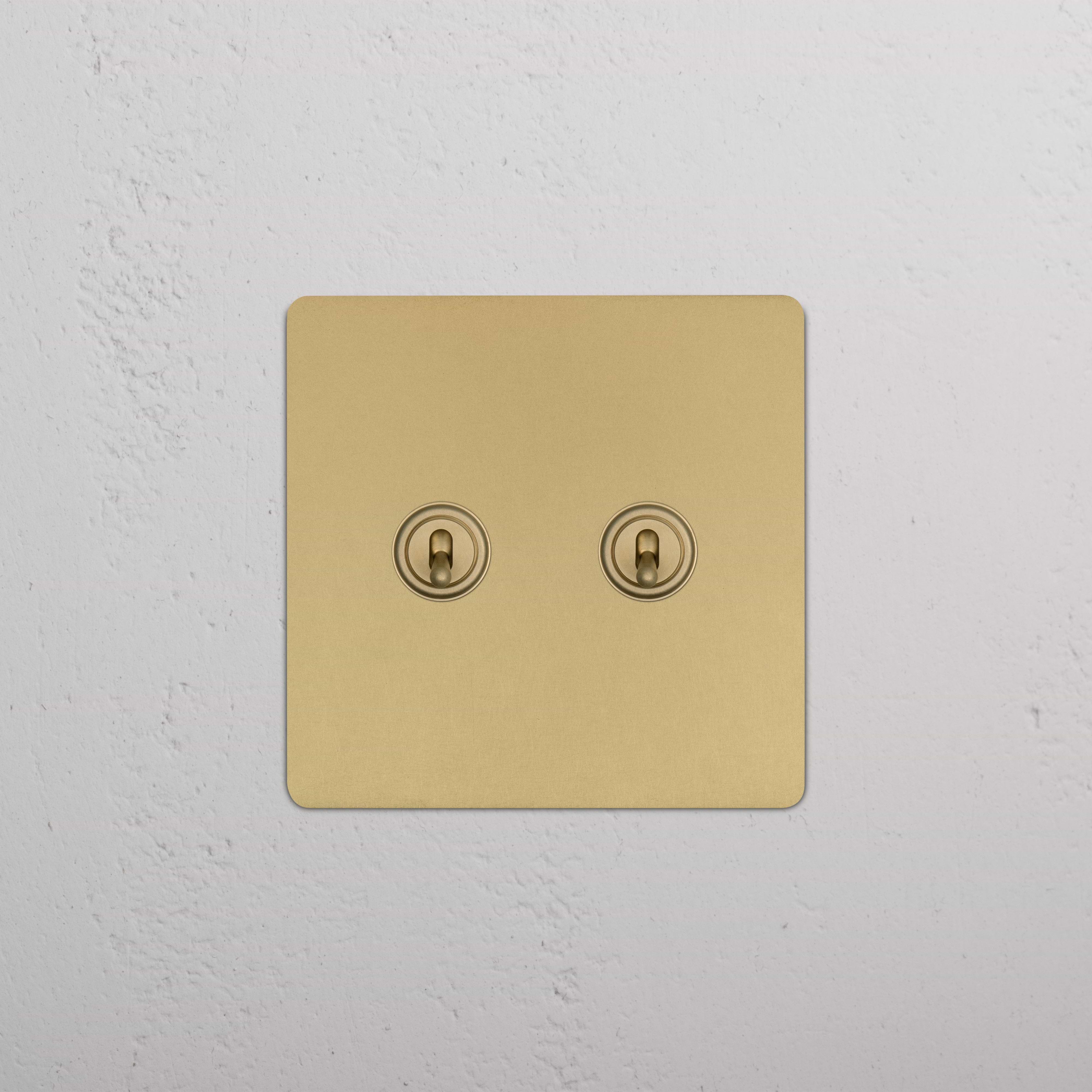 Kew 2G Toggle Switch in Antique Brass with round toggles, shown installed on a white textured wall.