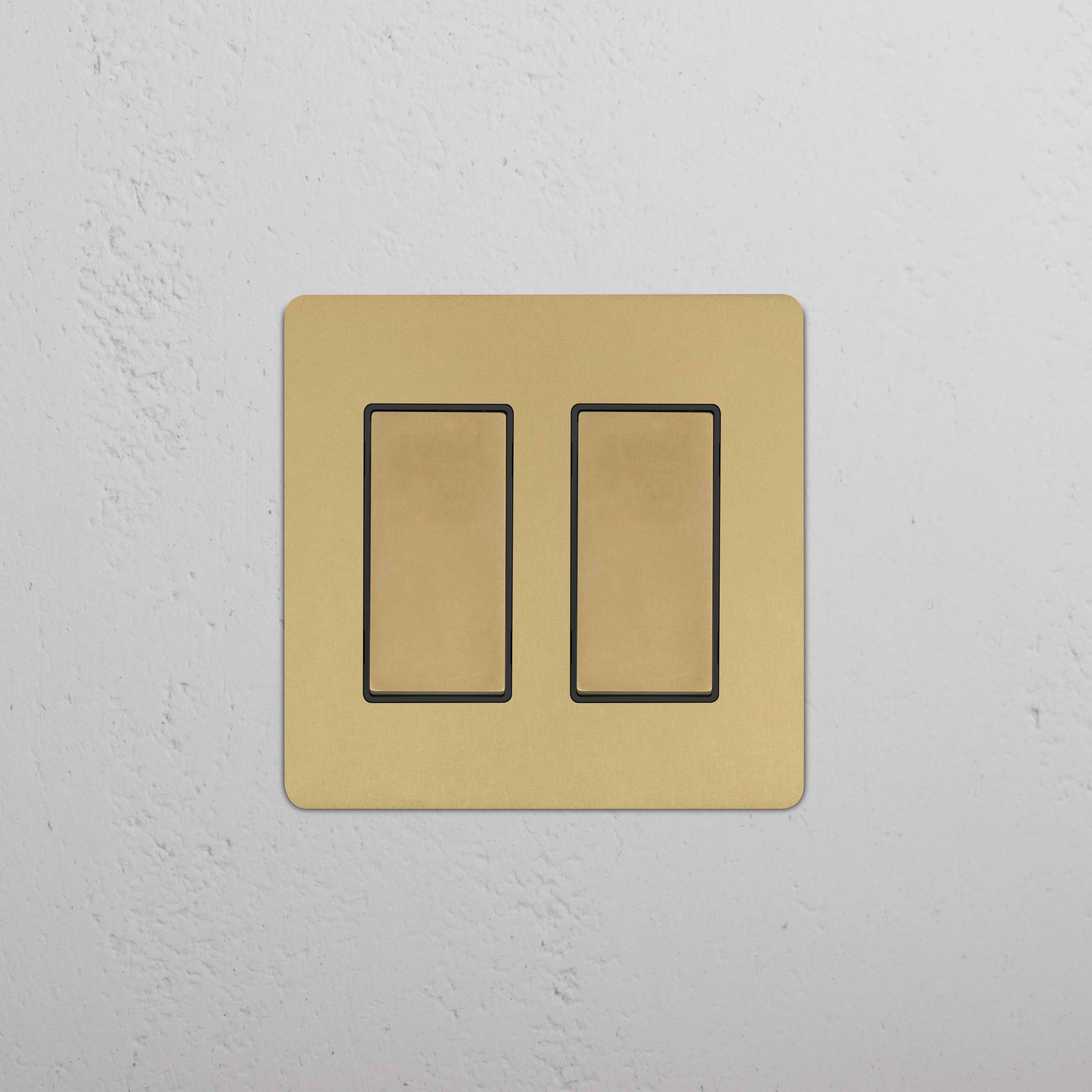 Kew 2G Rocker Switch in Antique Brass Black with two black rocker buttons, shown mounted on a wall.