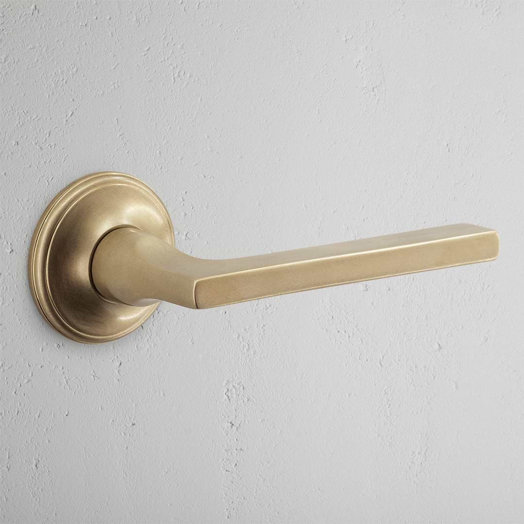 Clayton Passage Door Handle Set in antique brass with round base and straight lever on a light gray wall.