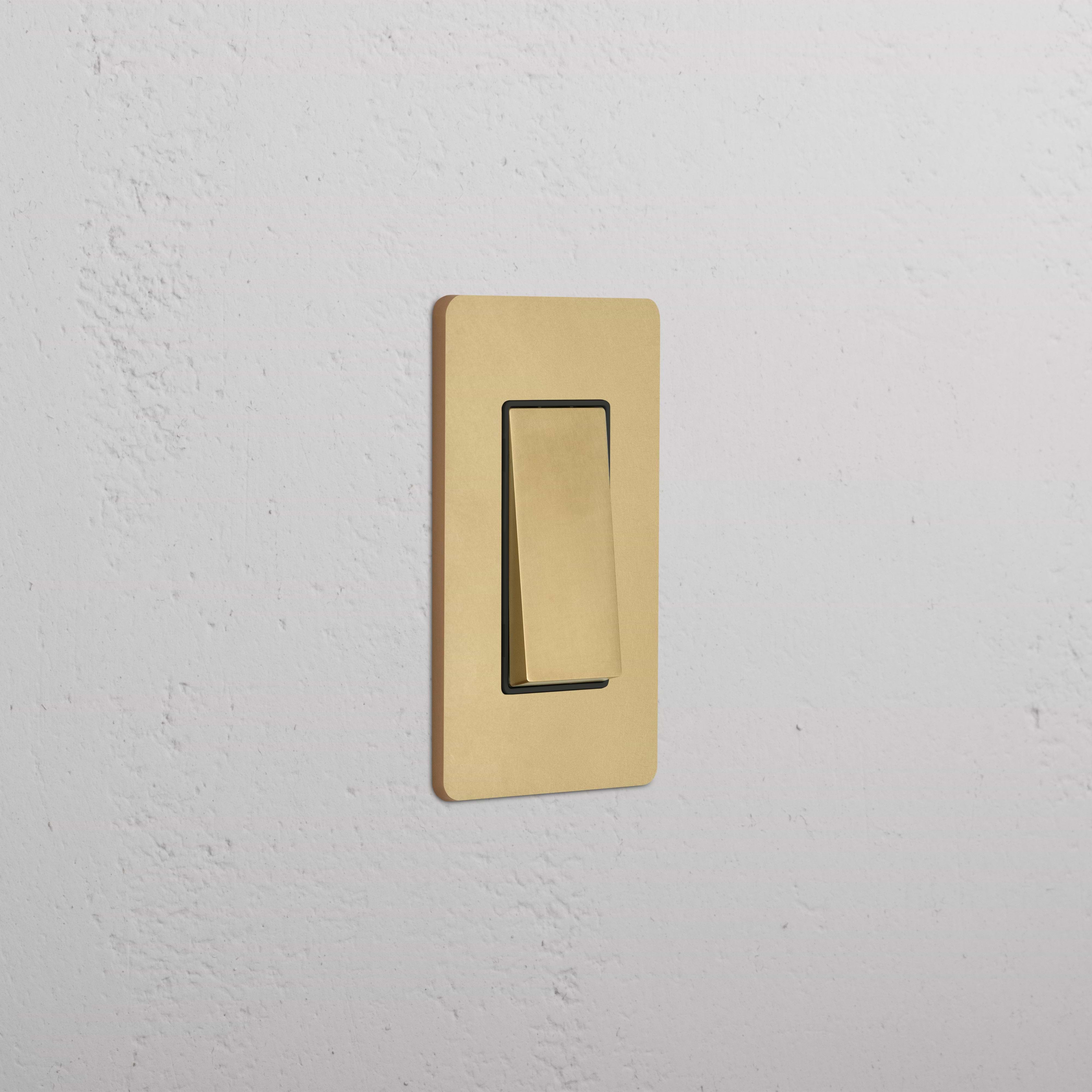 Grove 1G Rocker Switch in Antique Brass Black with a gold-tone rectangular plate on a white textured wall.