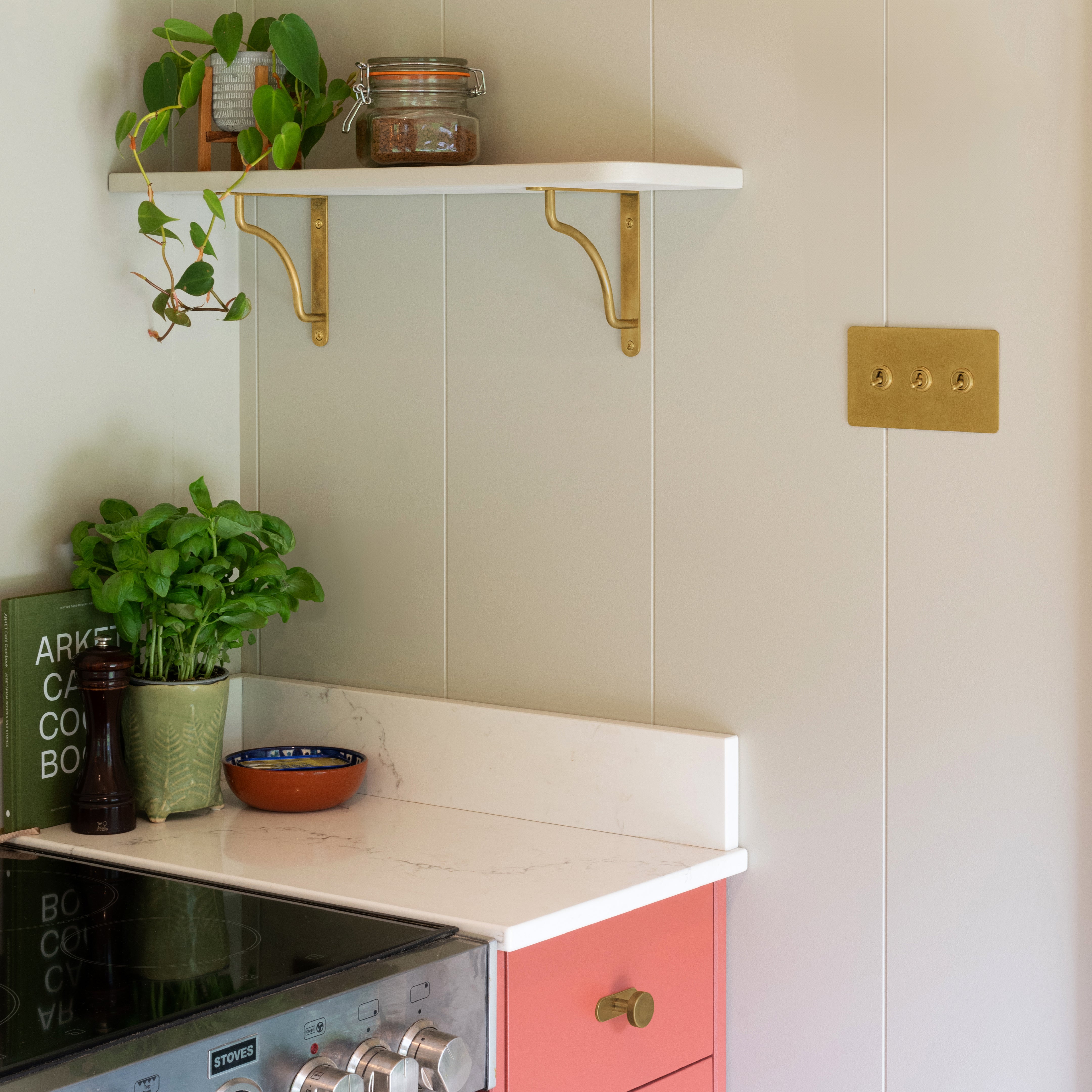 Small kitchen corner: pink cabinets, white shelf with Corston Antique Brass Kilburn Brackets holding jars, a plant, basil pot, and stove oven.