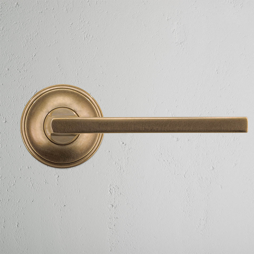 Clayton Passage Door Handle Set in antique brass with round base and straight lever on a gray wall.