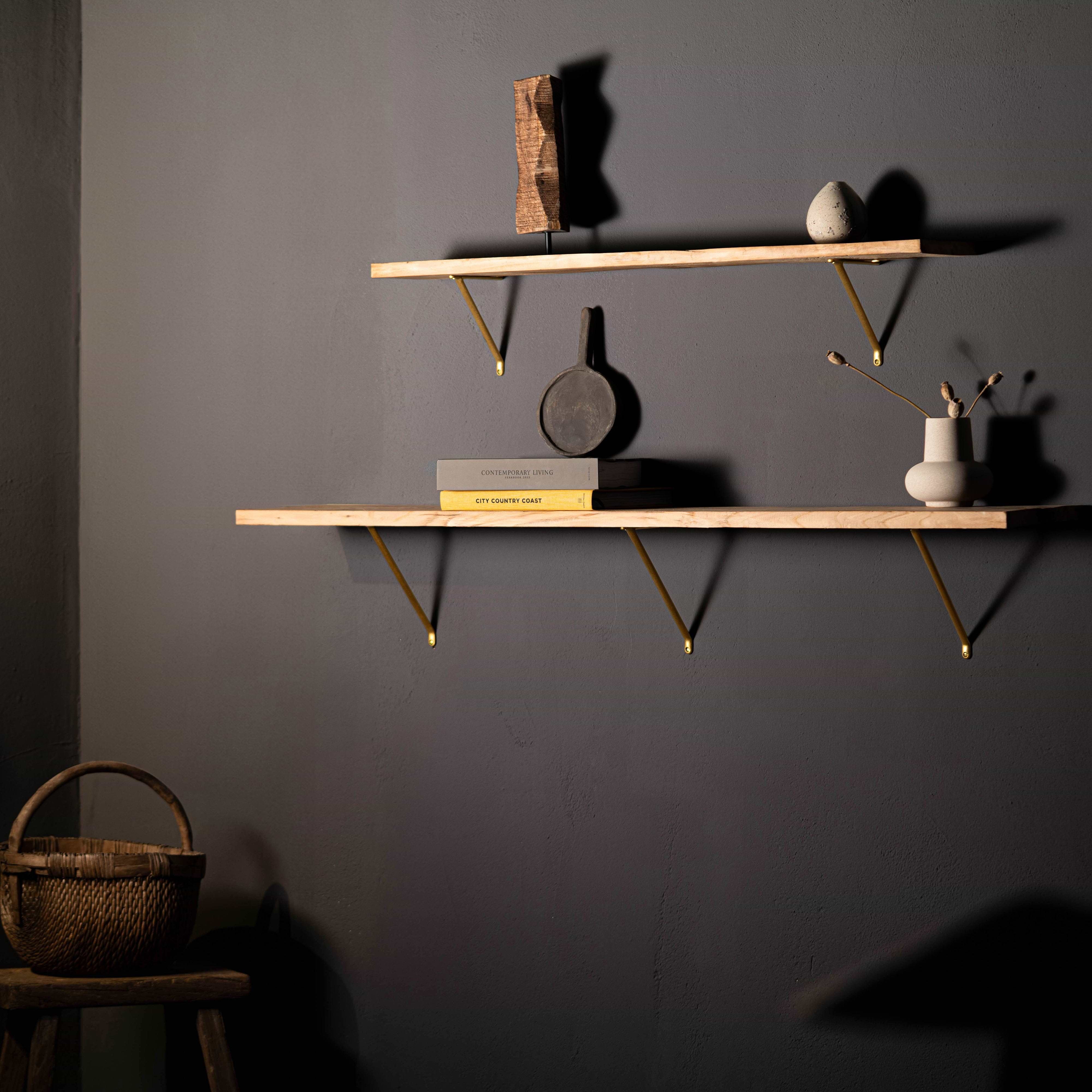 Small Brixton Shelf Bracket in Antique Brass supports wooden shelves holding minimalist decor on a dark gray wall.