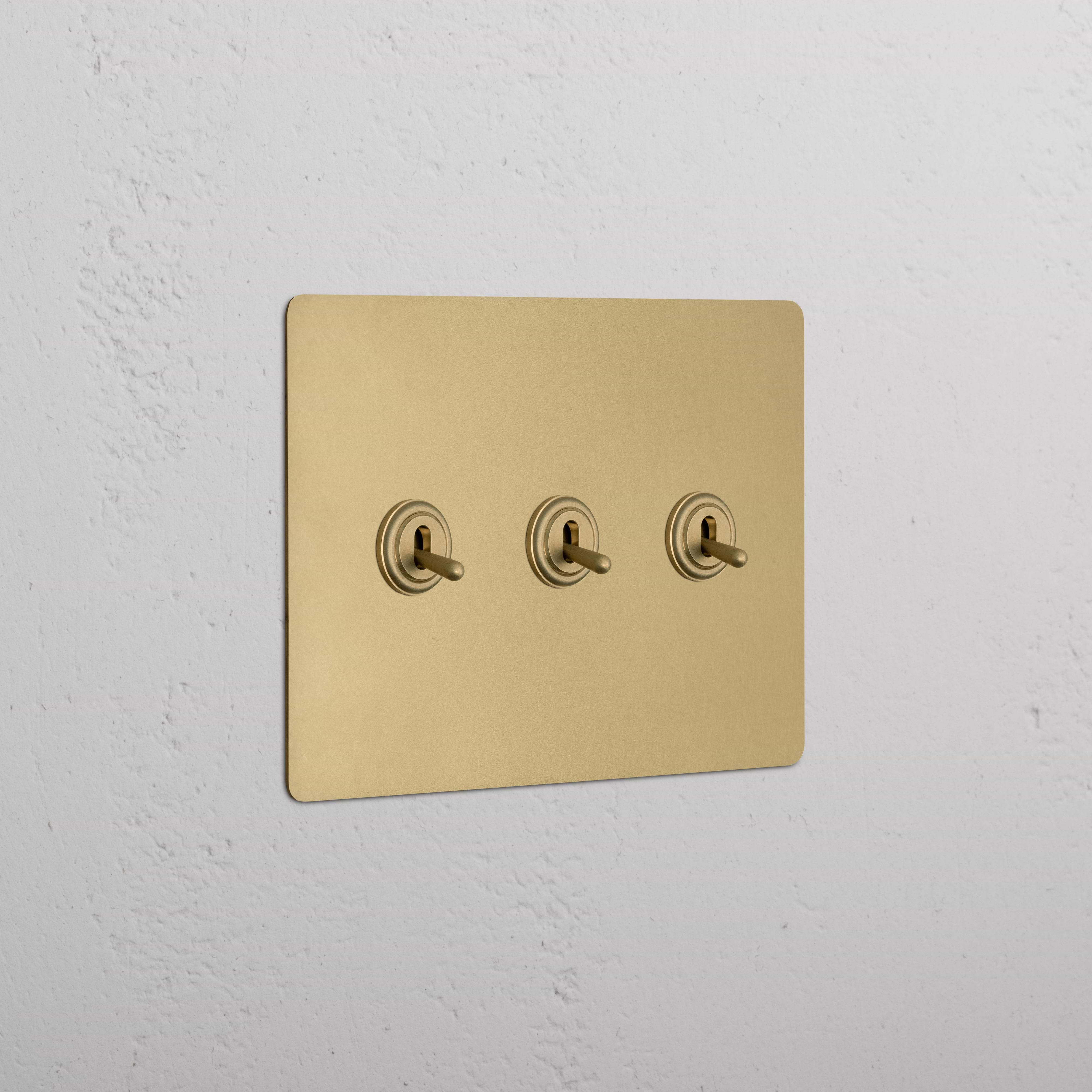 Kew 3G Toggle Switch in antique brass finish with three centered toggles on a white textured wall.