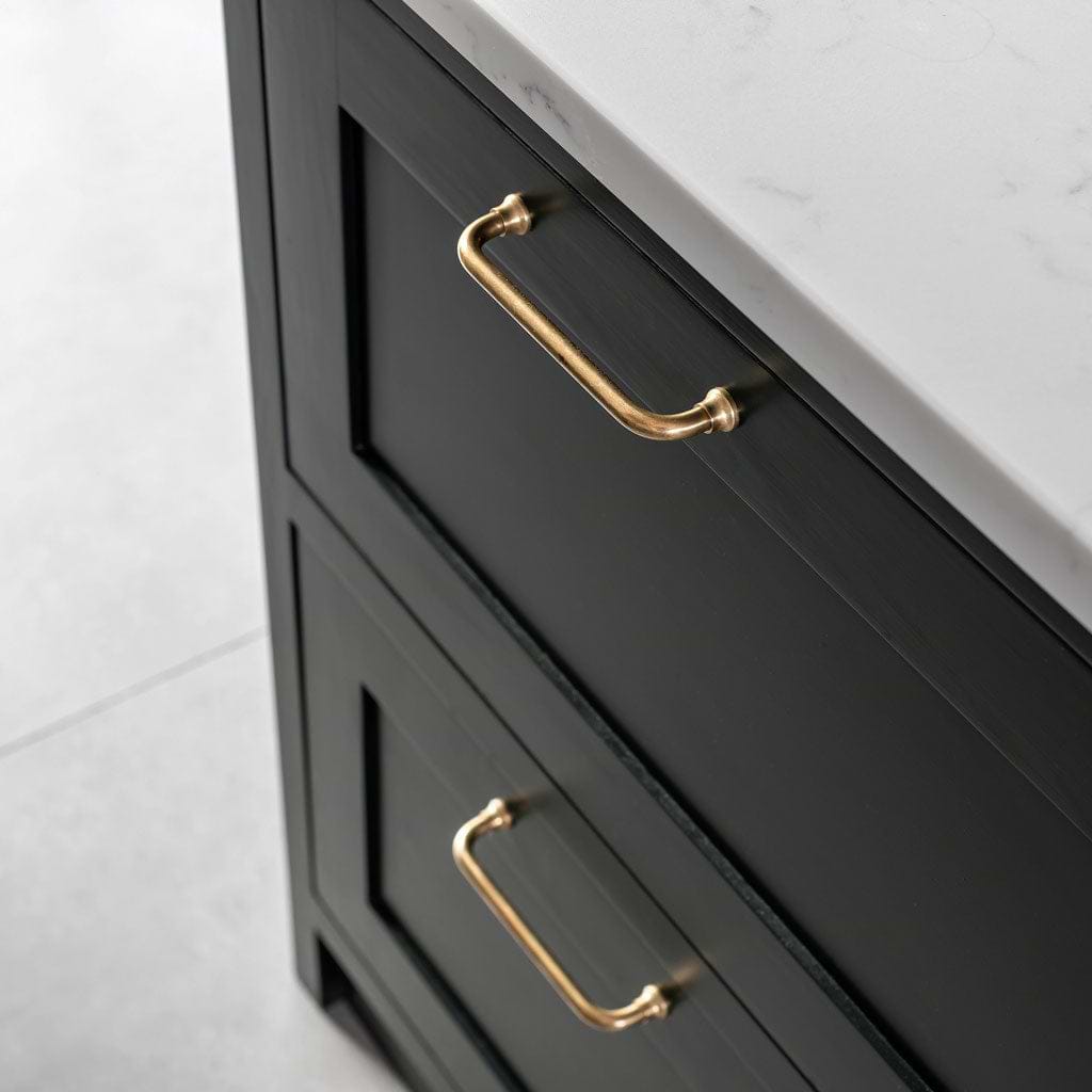 Small Antique Brass Sycamore Furniture Handle fitted on dark cabinet drawers beneath a white marble countertop.