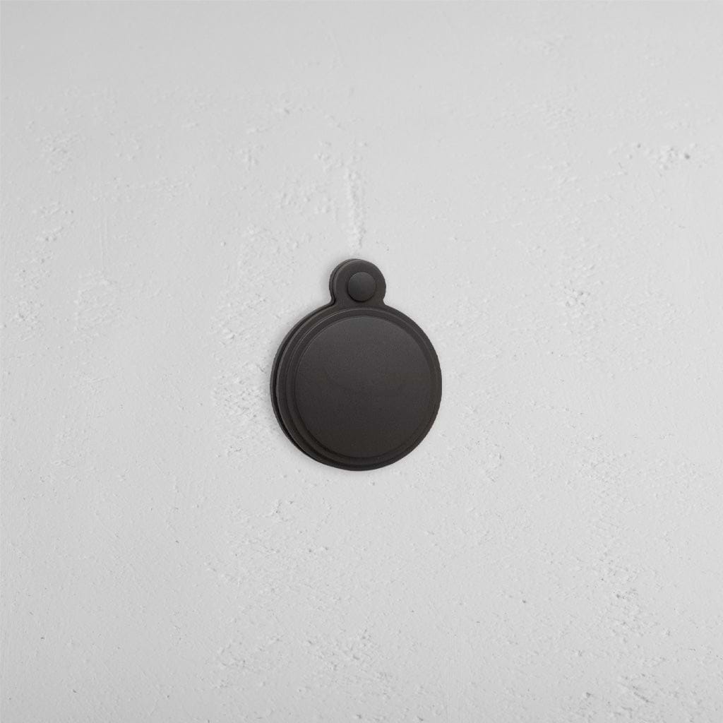 Bronze Warwick Covered Key Escutcheon, round with cover and small loop, mounted on a white textured wall.
