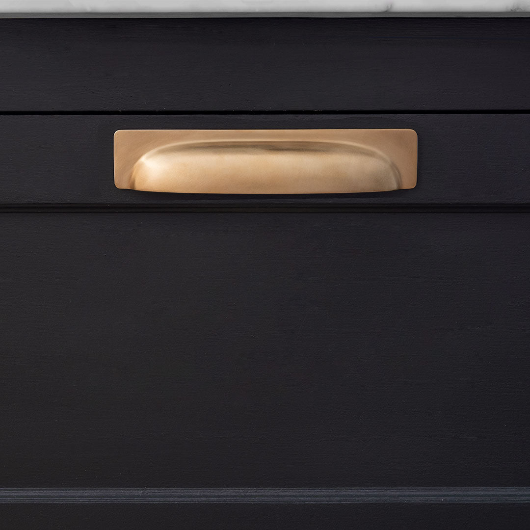 A medium antique brass Elm cup handle mounted on a black cabinet drawer under a white countertop.