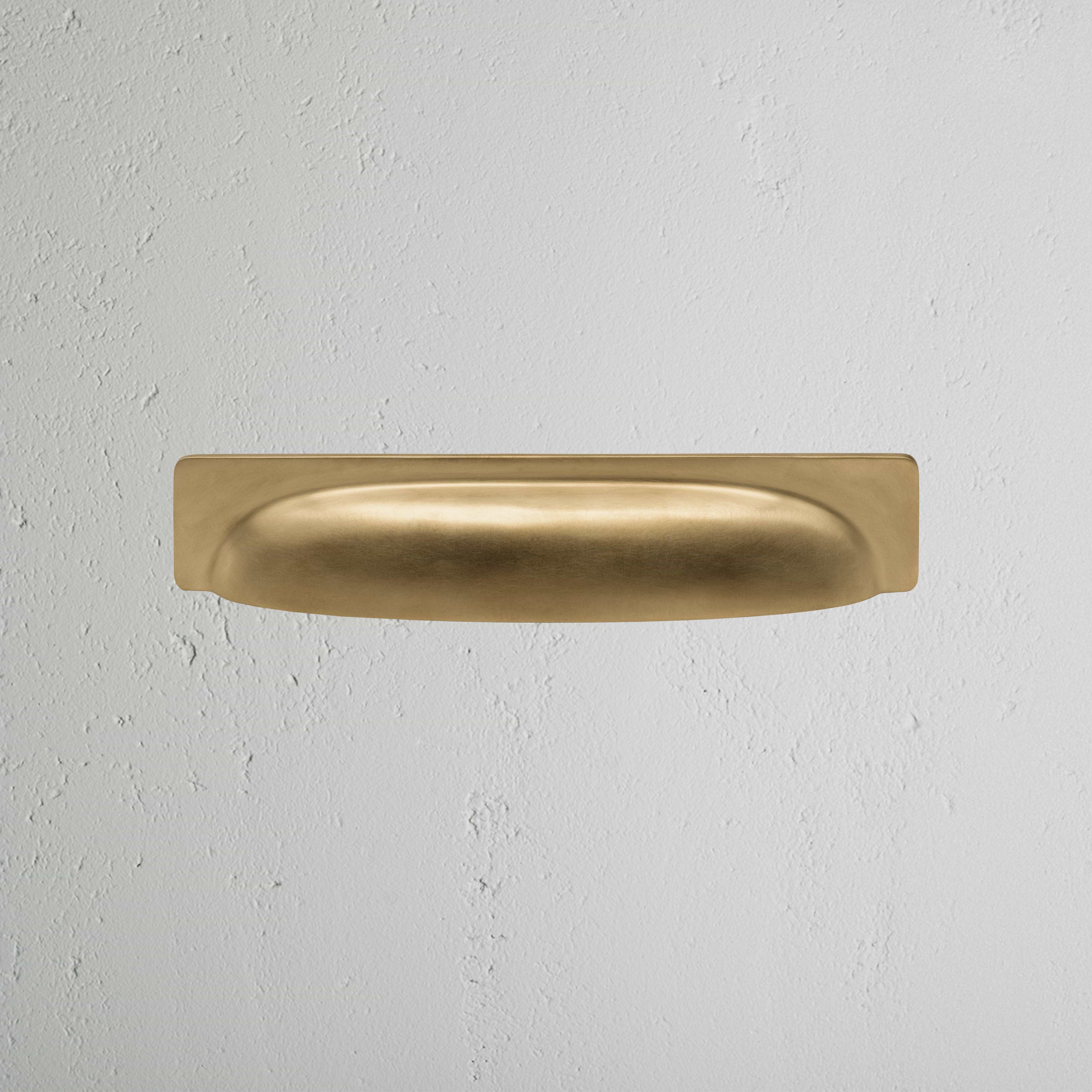 Elm Cup Handle Medium in antique brass finish, featuring a smooth curved design on a light gray surface.