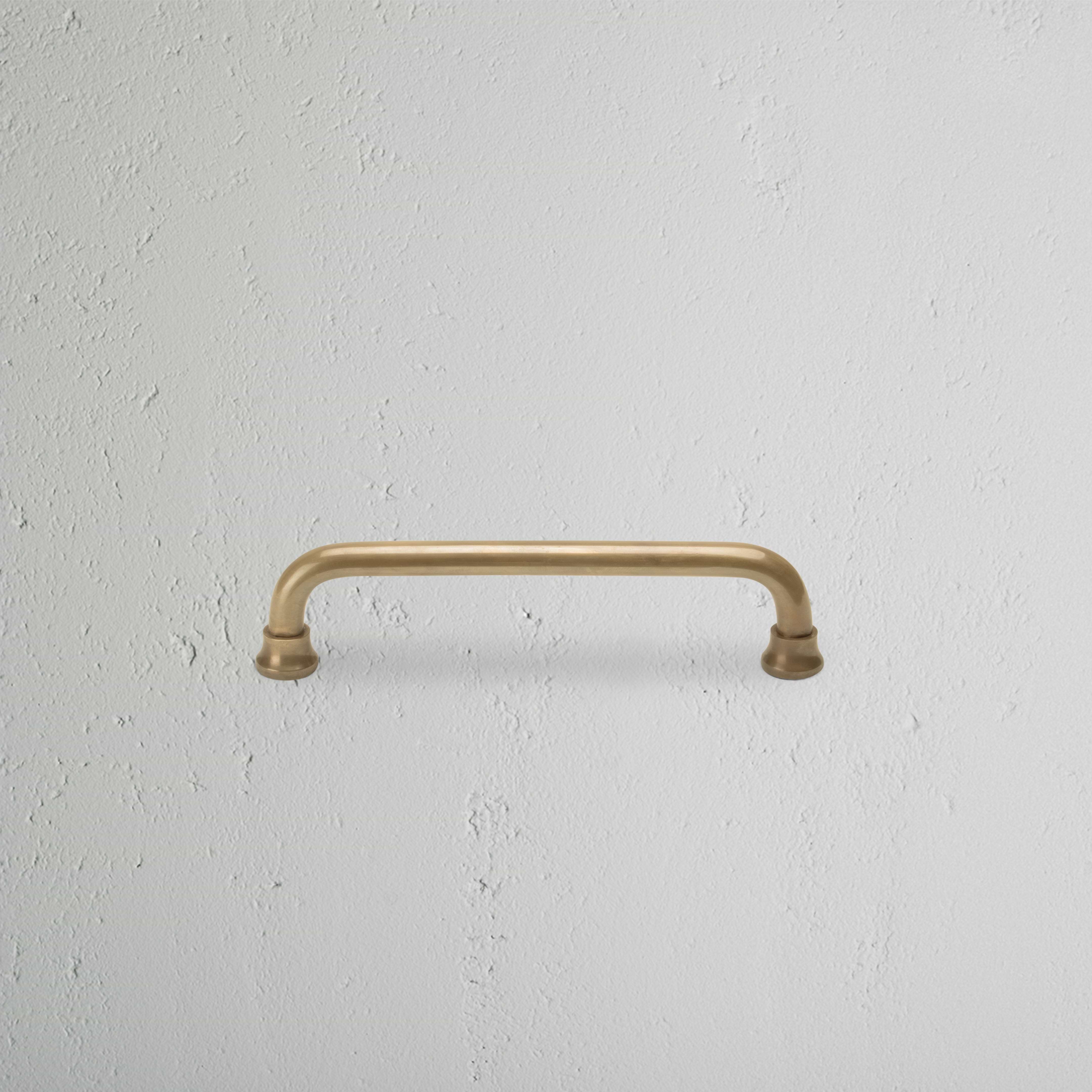 Sycamore Furniture Handle Small in antique brass finish, shown mounted on a white textured surface.