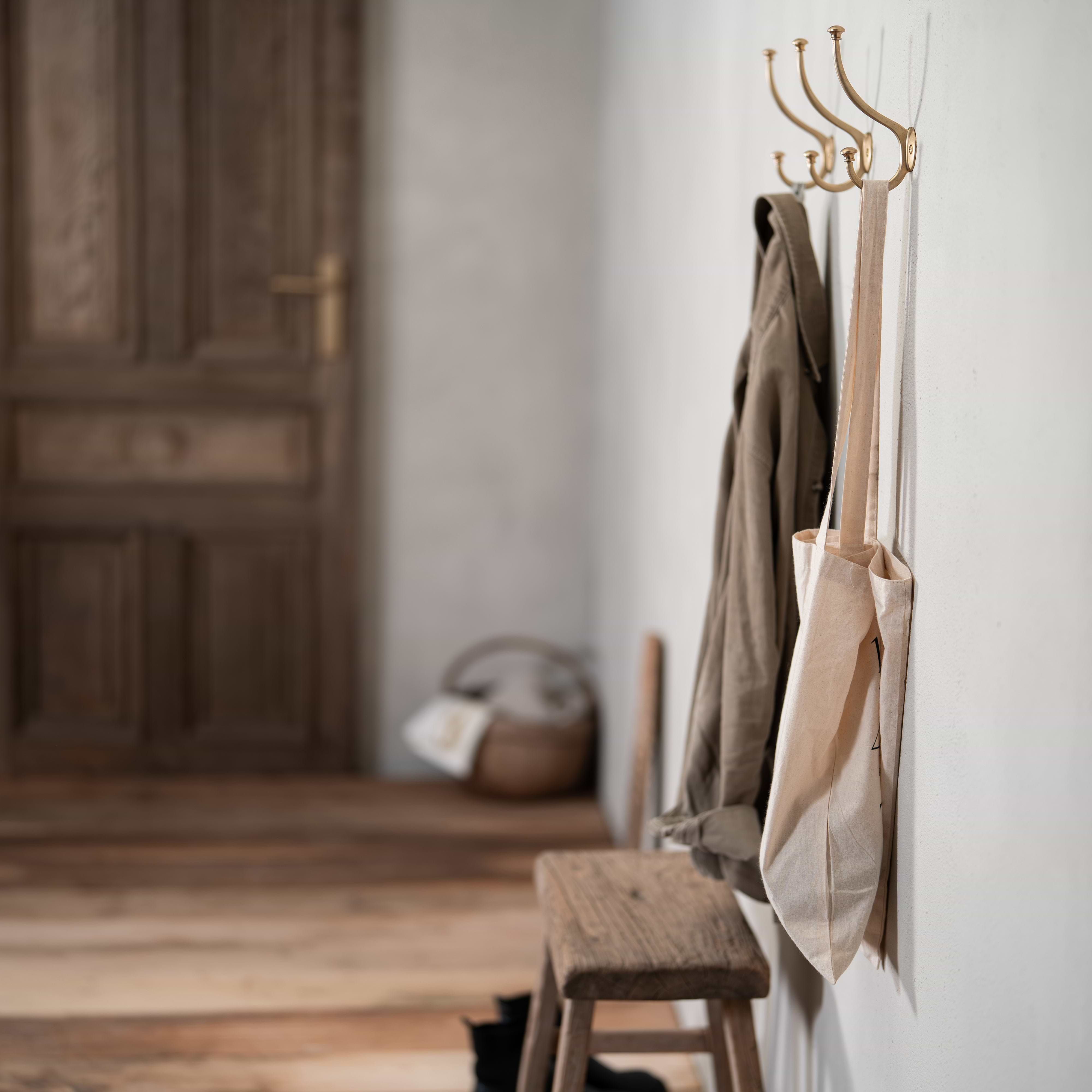 Antique brass Mews Hook mounted on a wall, holding a beige tote bag and a khaki coat.