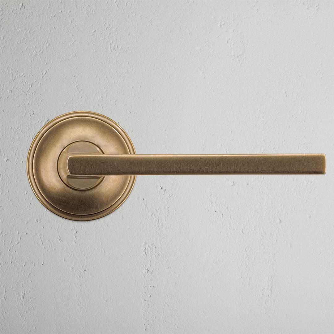 Clayton Passage Door Handle Set in antique brass with round base and straight lever on a gray wall.