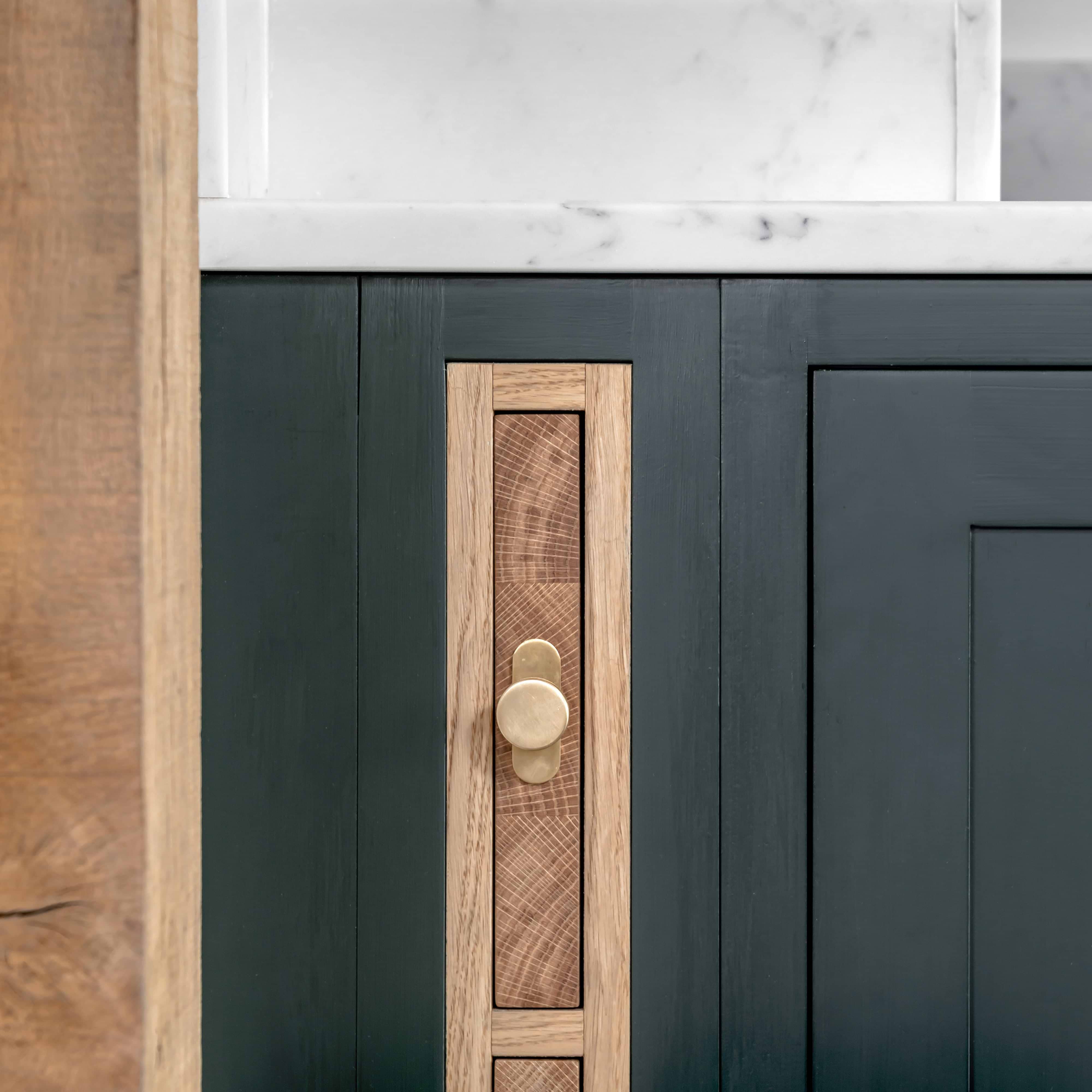 Close-up of a polished Kilburn Furniture Knob mounted on a dark green cabinet door with wood inlay.