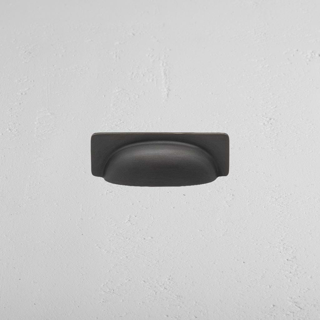Elm Cup Handle Small in bronze finish, cup-style drawer pull mounted on a white textured surface.