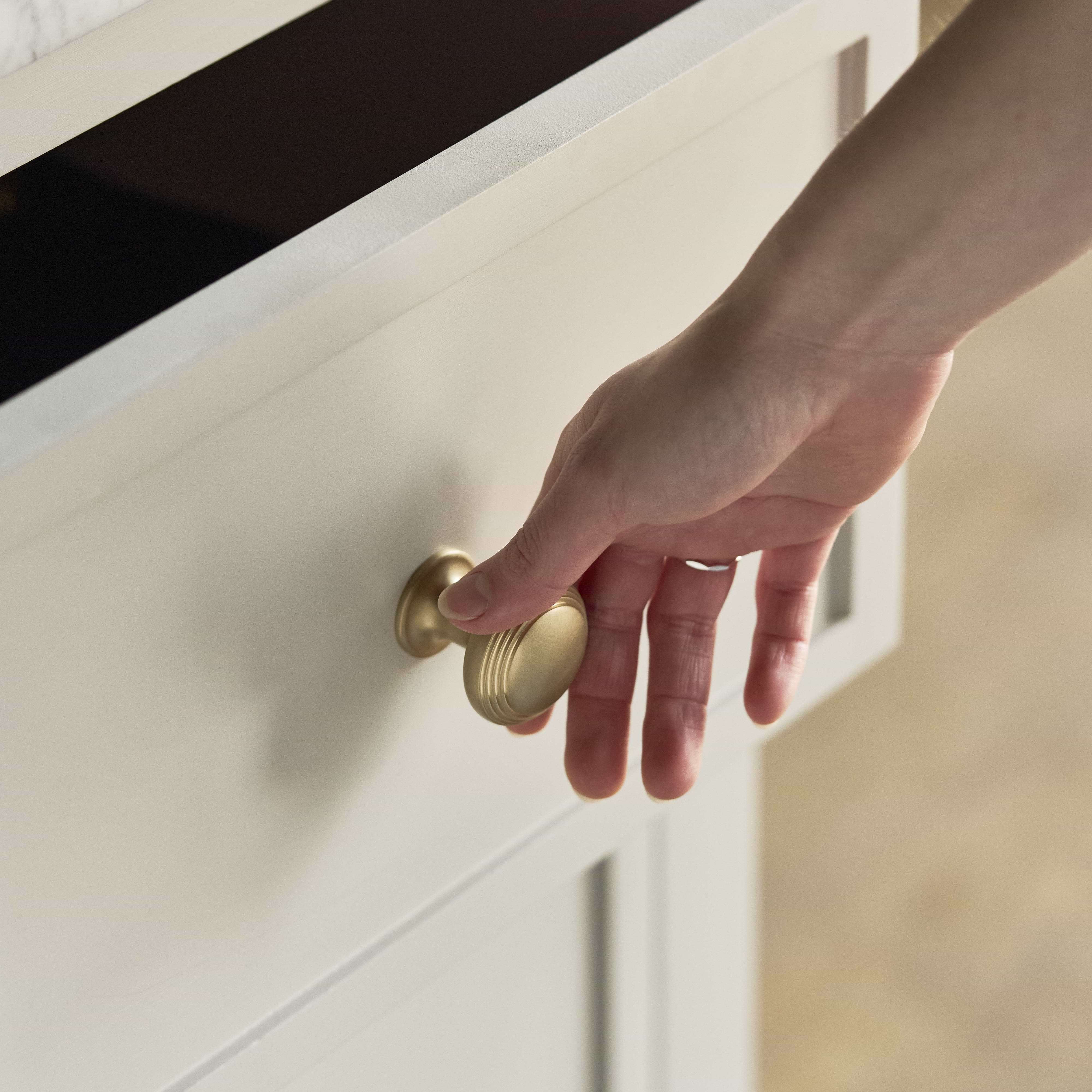 A hand grasps a Barlow Furniture Knob Medium in Antique Brass, attached to a light wood drawer.