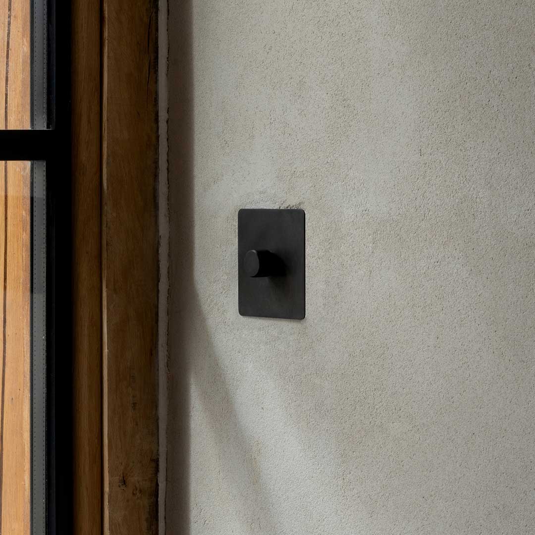 Grove 1G Dimming Retractive Toggle Switch in bronze, shown installed on a light gray wall.