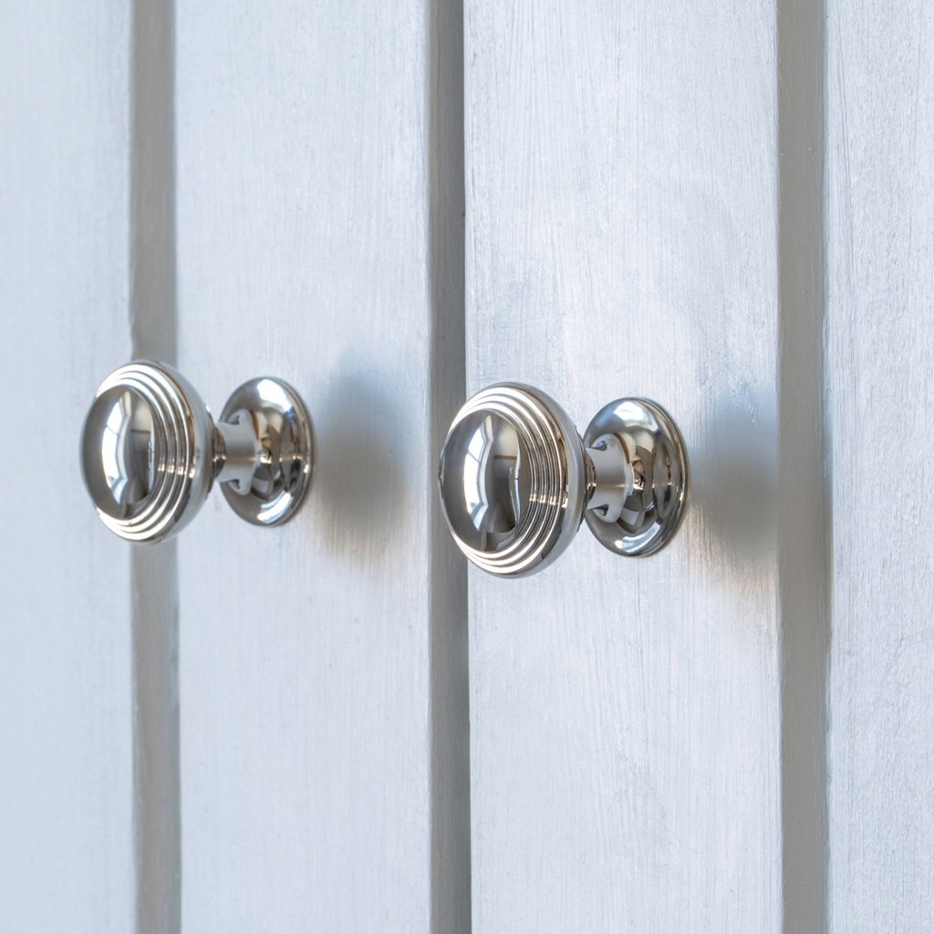Two small Barlow Furniture Knobs in polished nickel with ridged design on a light gray paneled surface.