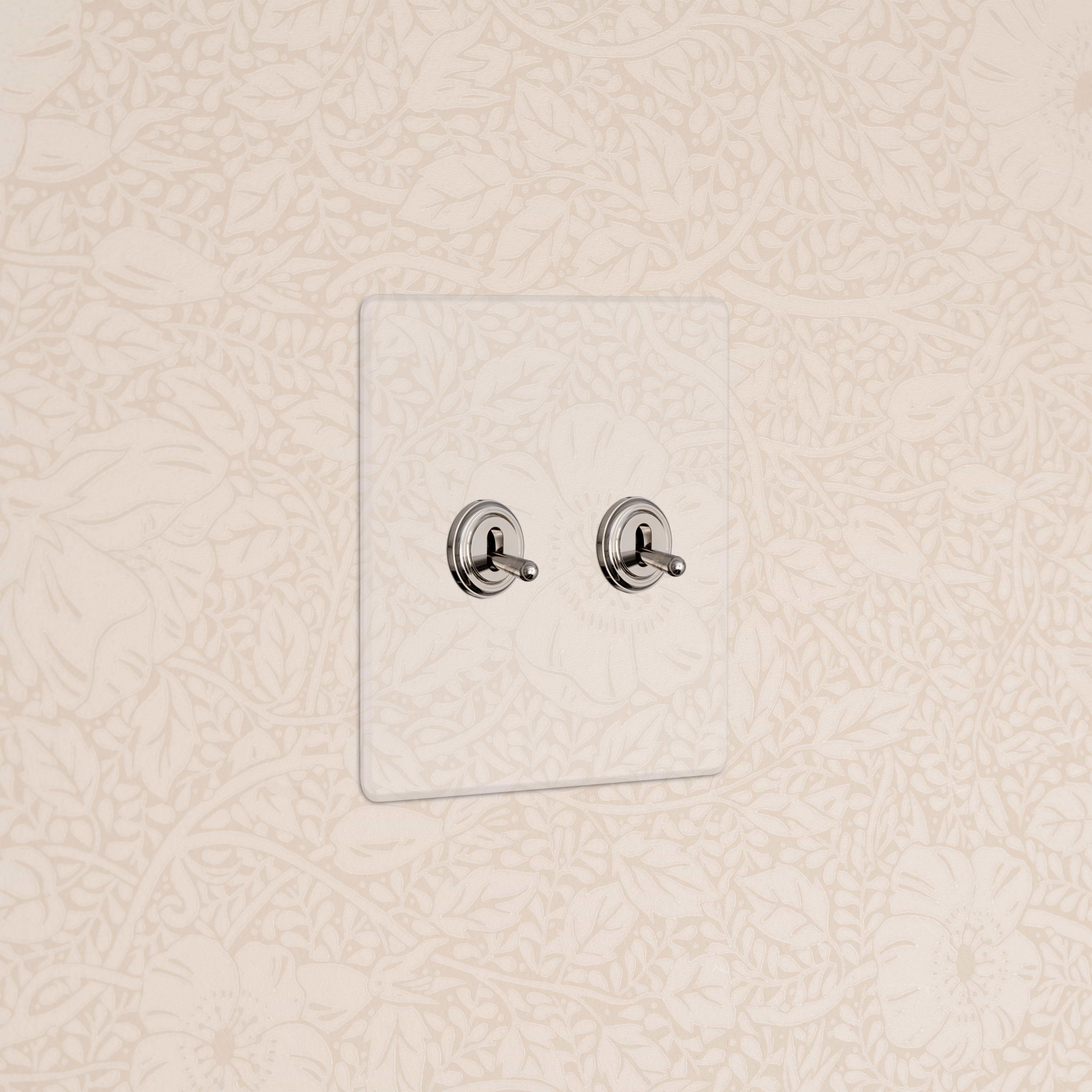 Kew 2G Toggle Switch in clear polished nickel with two toggles on a white wall plate against beige floral wallpaper.