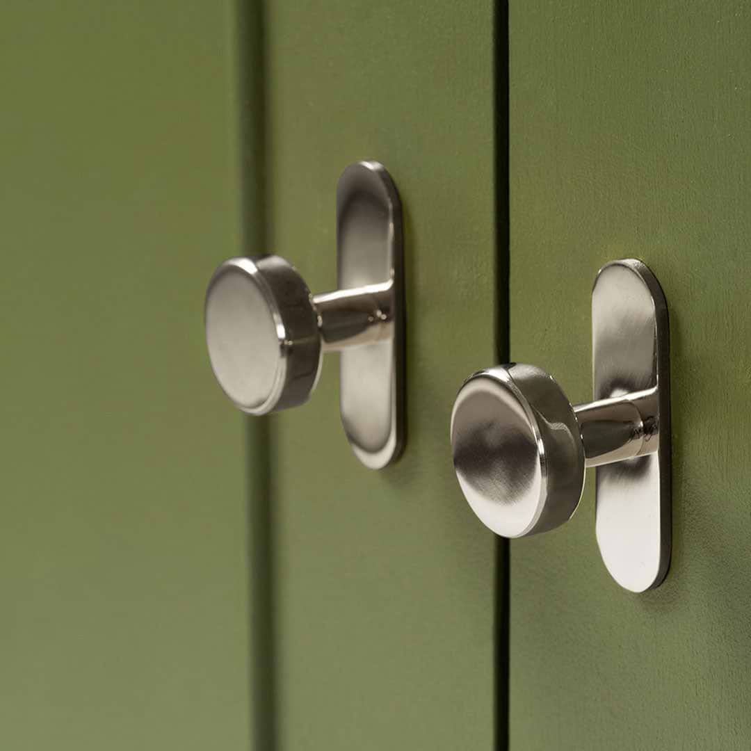 Two polished nickel Kilburn Furniture Knobs mounted on green cabinet doors, shown in soft lighting.